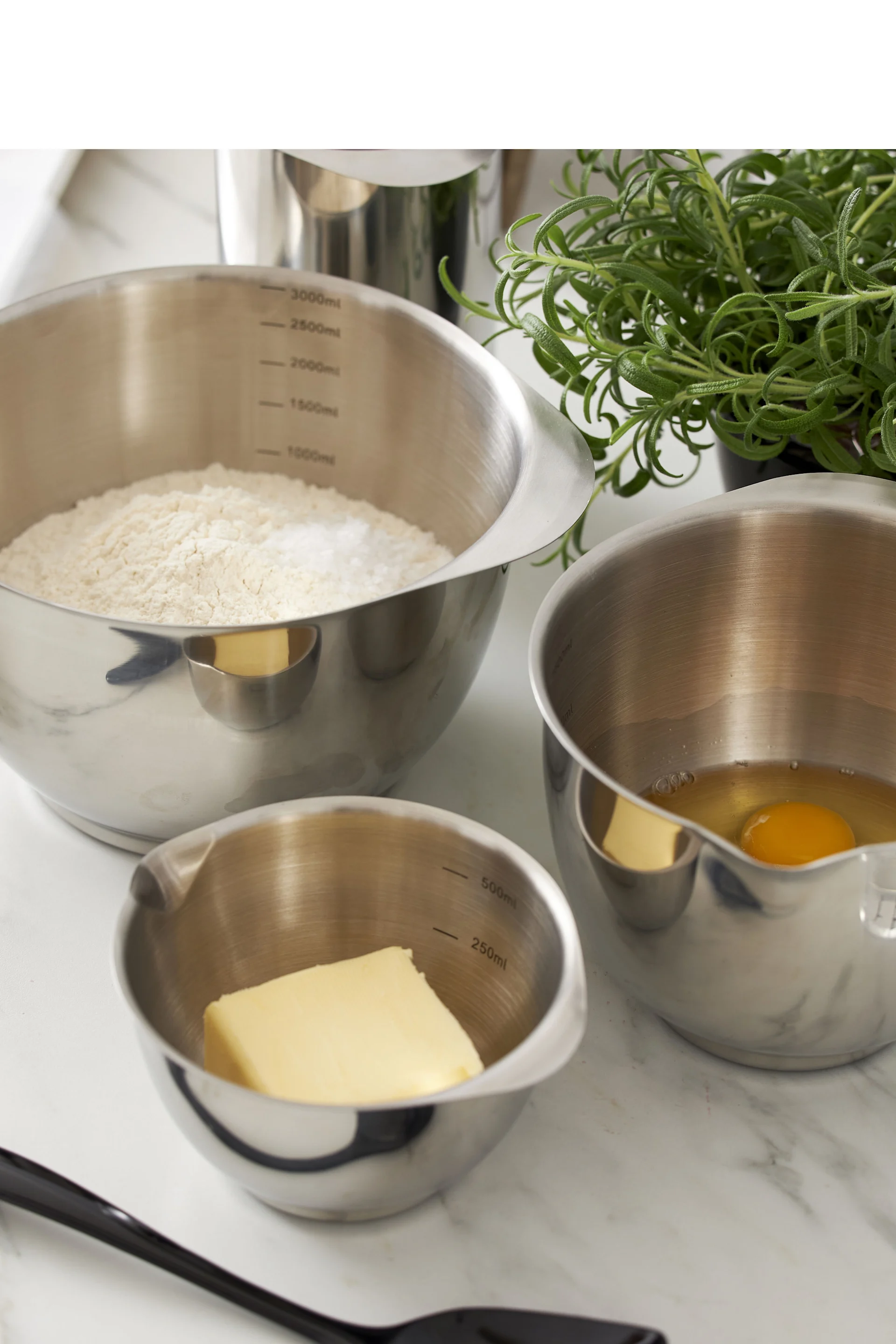 Rührschüsseln aus Edelstahl mit Mehl, Ei, Butter und einer Pflanze auf einer Marmorplatte.