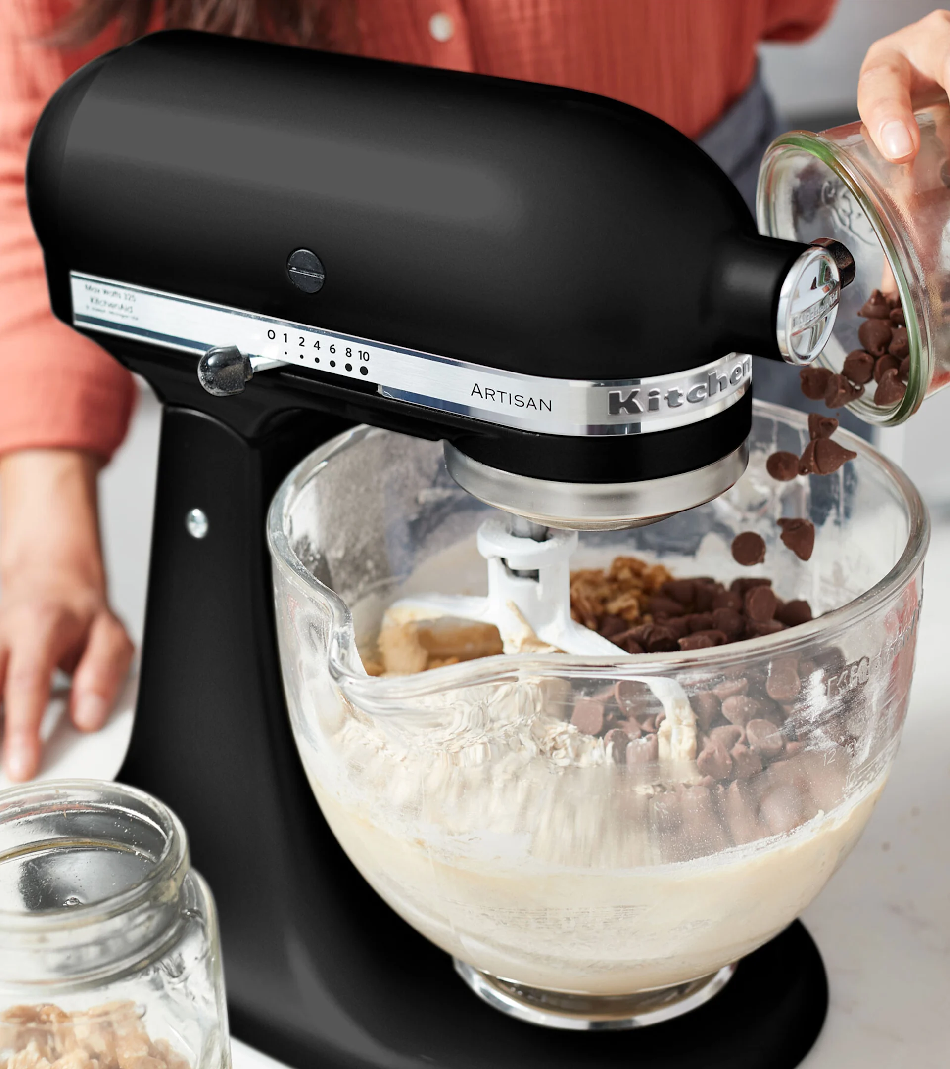 Eine schwarze Küchenmaschine mit einer klaren Glasschüssel. Schokoladenstückchen werden in die Schüssel gegossen, die Teig und andere Zutaten enthält.
