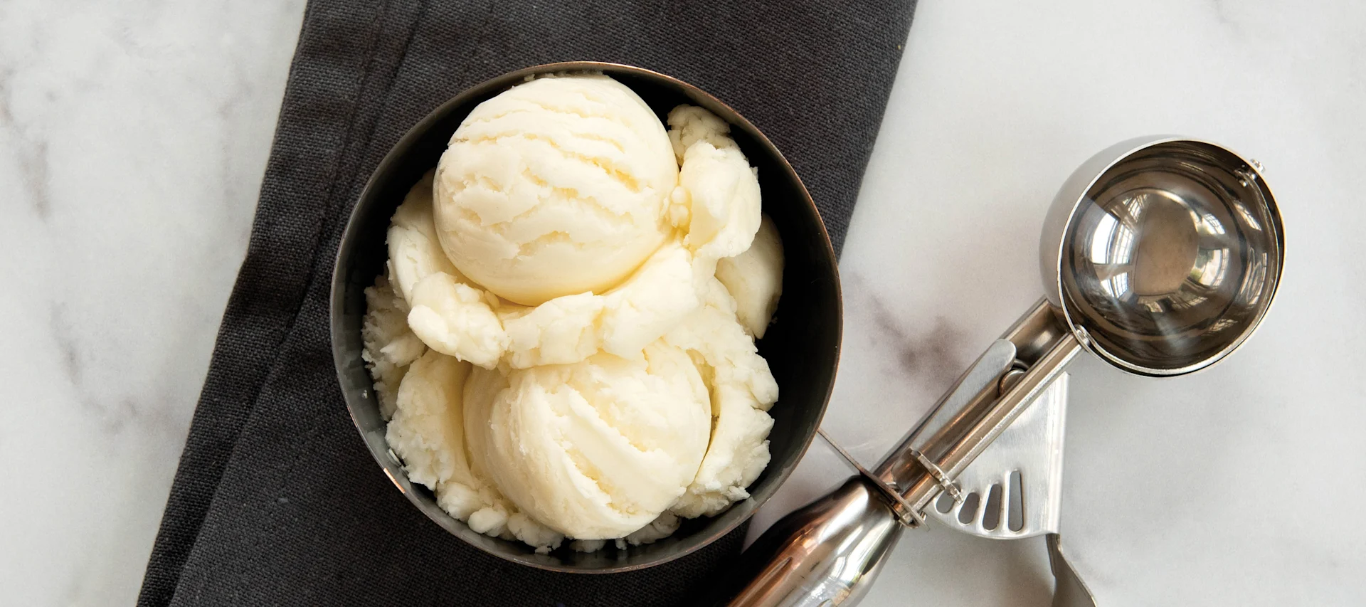Kugeln Vanilleeis in einer dunklen Schüssel neben einem Eislöffel auf einer Marmoroberfläche.