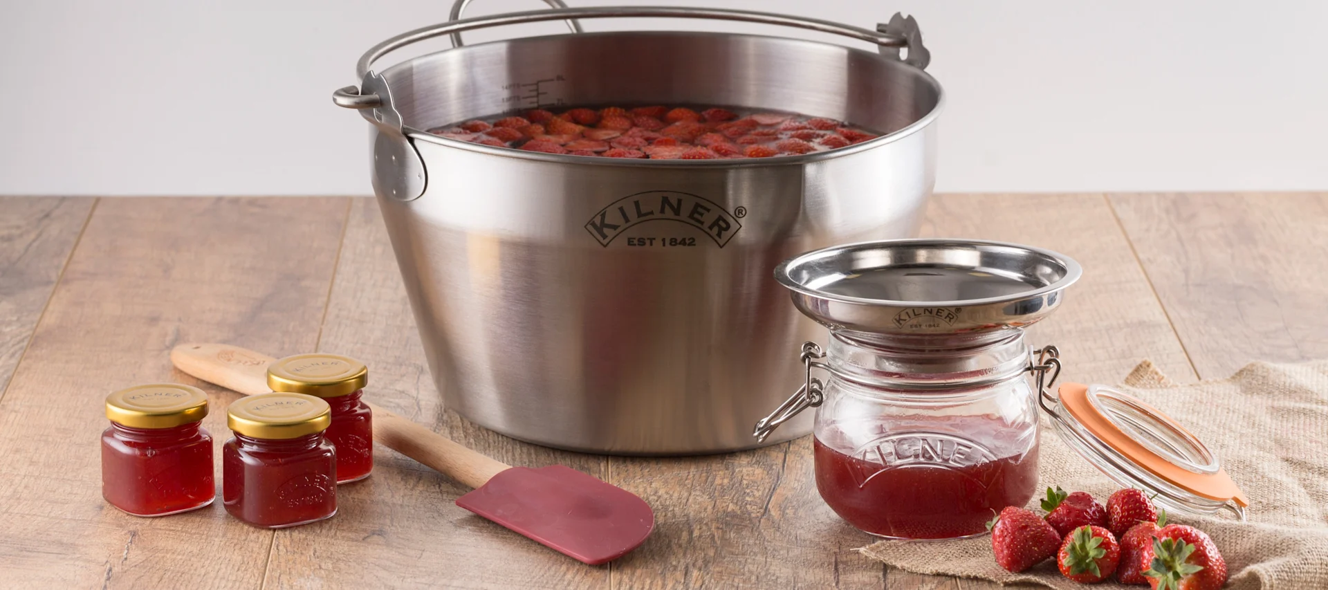 Edelstahl-Einkochtopf gefüllt mit Erdbeeren, zwei kleine Marmeladengläser, ein leeres Glas mit Trichter, ein roter Spatel und frische Erdbeeren auf einem Holztisch.