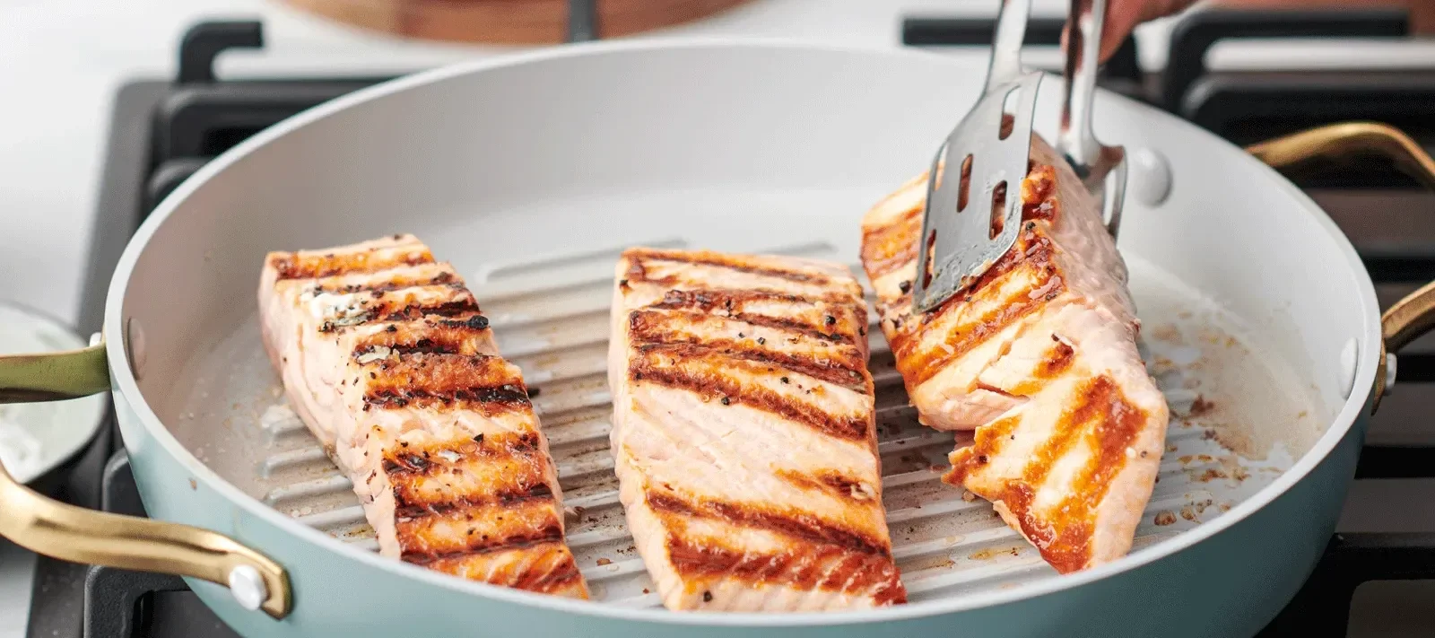 In einer blauen Pfanne von Greenpan werden drei Stücke Lachs gebraten und eines wird soeben von einer Zange gewendet.