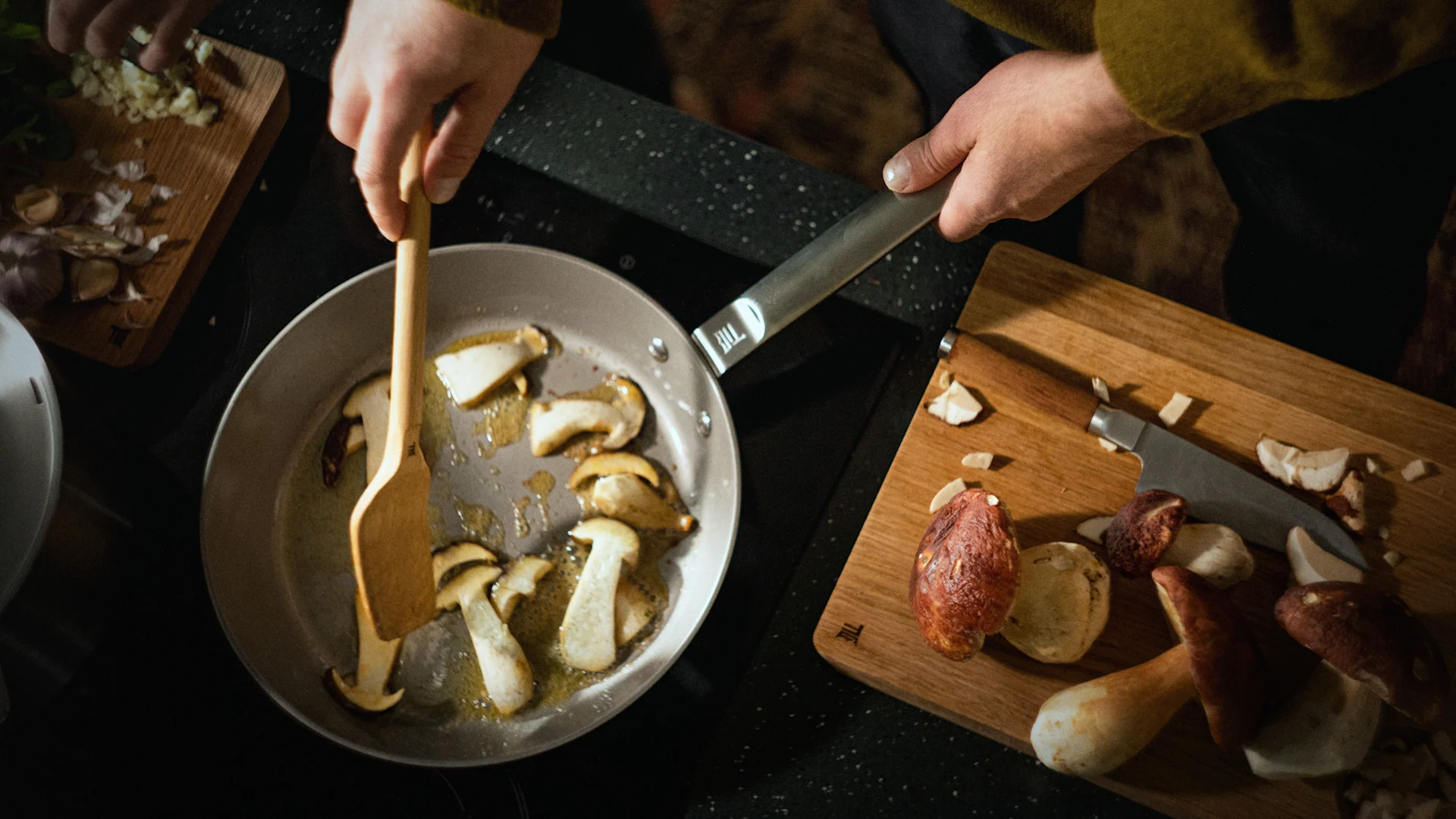 Hände braten Pilze in einer Pfanne, mit gehacktem Knoblauch und weiteren Pilzen auf Schneidebrettern.
