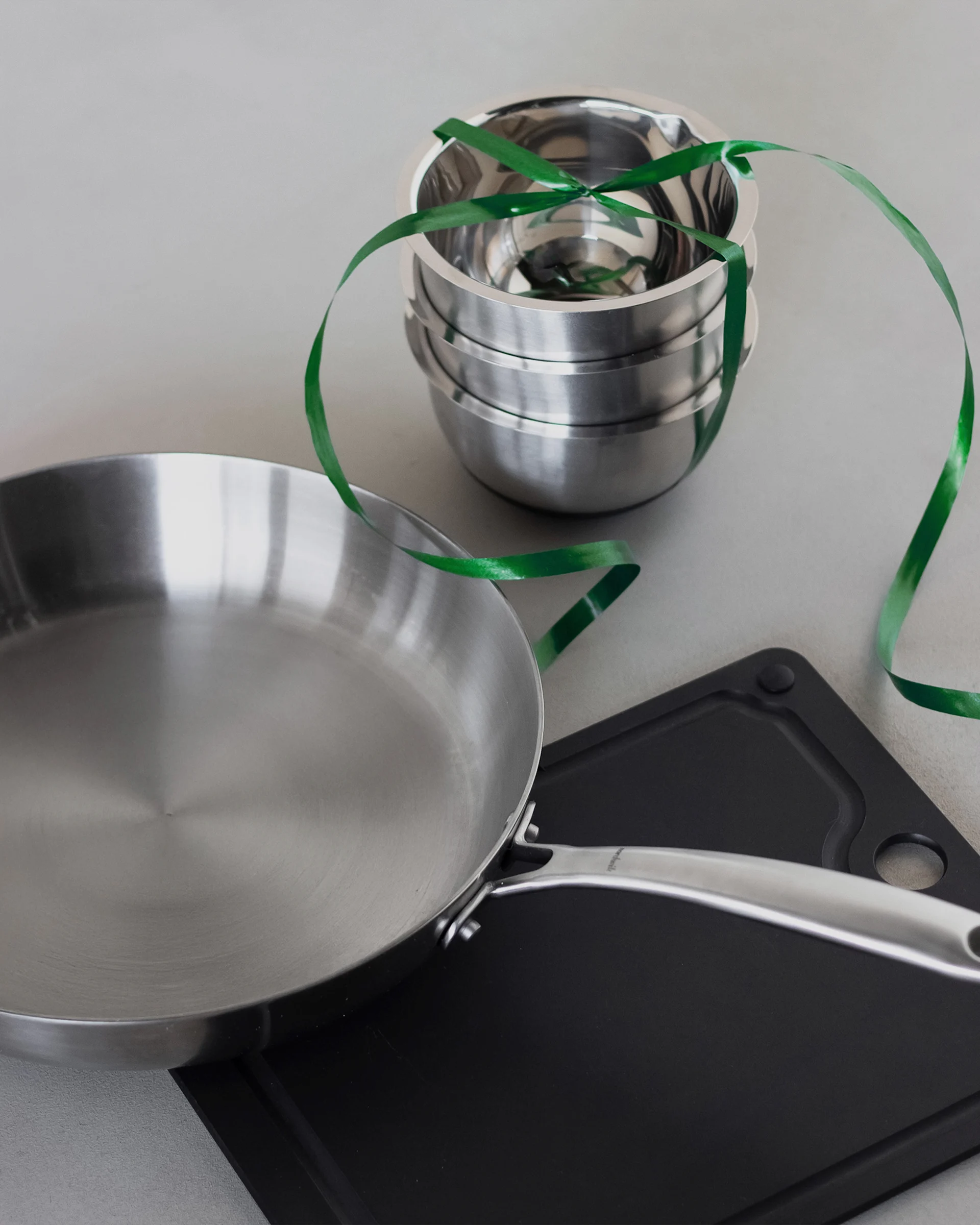 Eine Edelstahlpfanne, ein Stapel Metallschüsseln mit einem grünen Band und ein schwarzes Schneidebrett auf hellem Untergrund.