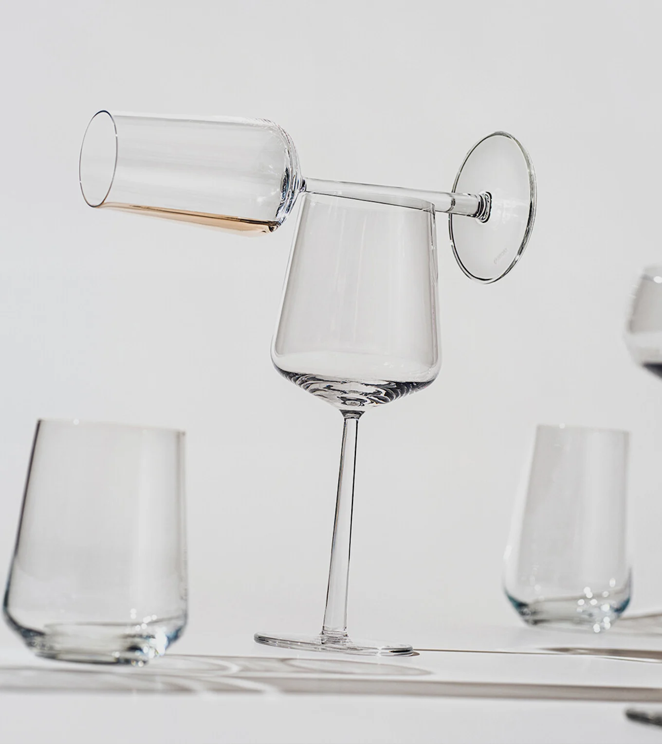 Eine Champagnerflöte mit bernsteinfarbener Flüssigkeit balanciert horizontal auf dem Rand eines aufrechten Weinglases, mit weiteren klaren Gläsern im Hintergrund.