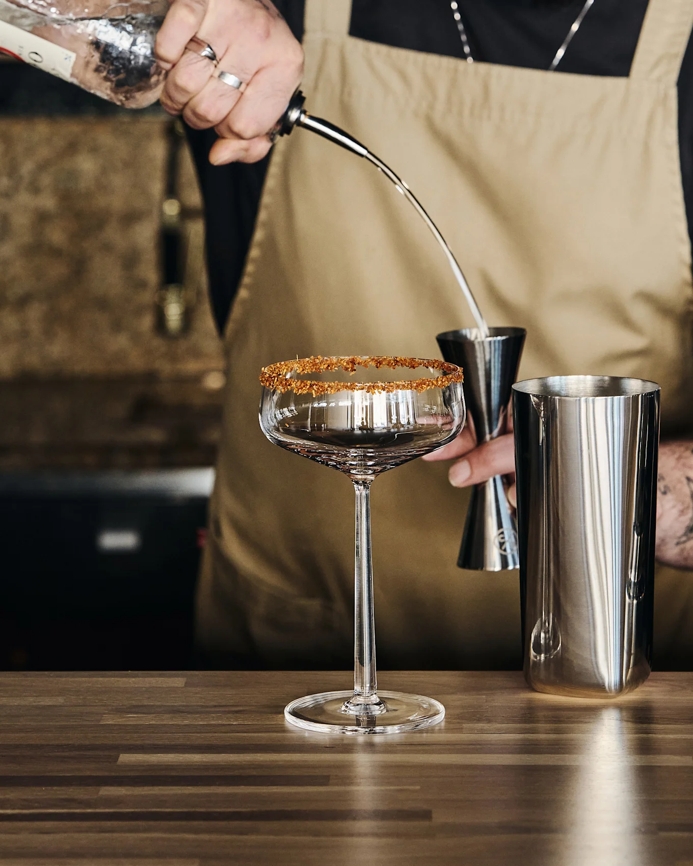 Barkeeper gießt Flüssigkeit aus einer Flasche in einen Jigger über einen Shaker, neben einem Stielglas mit Zuckerrand.