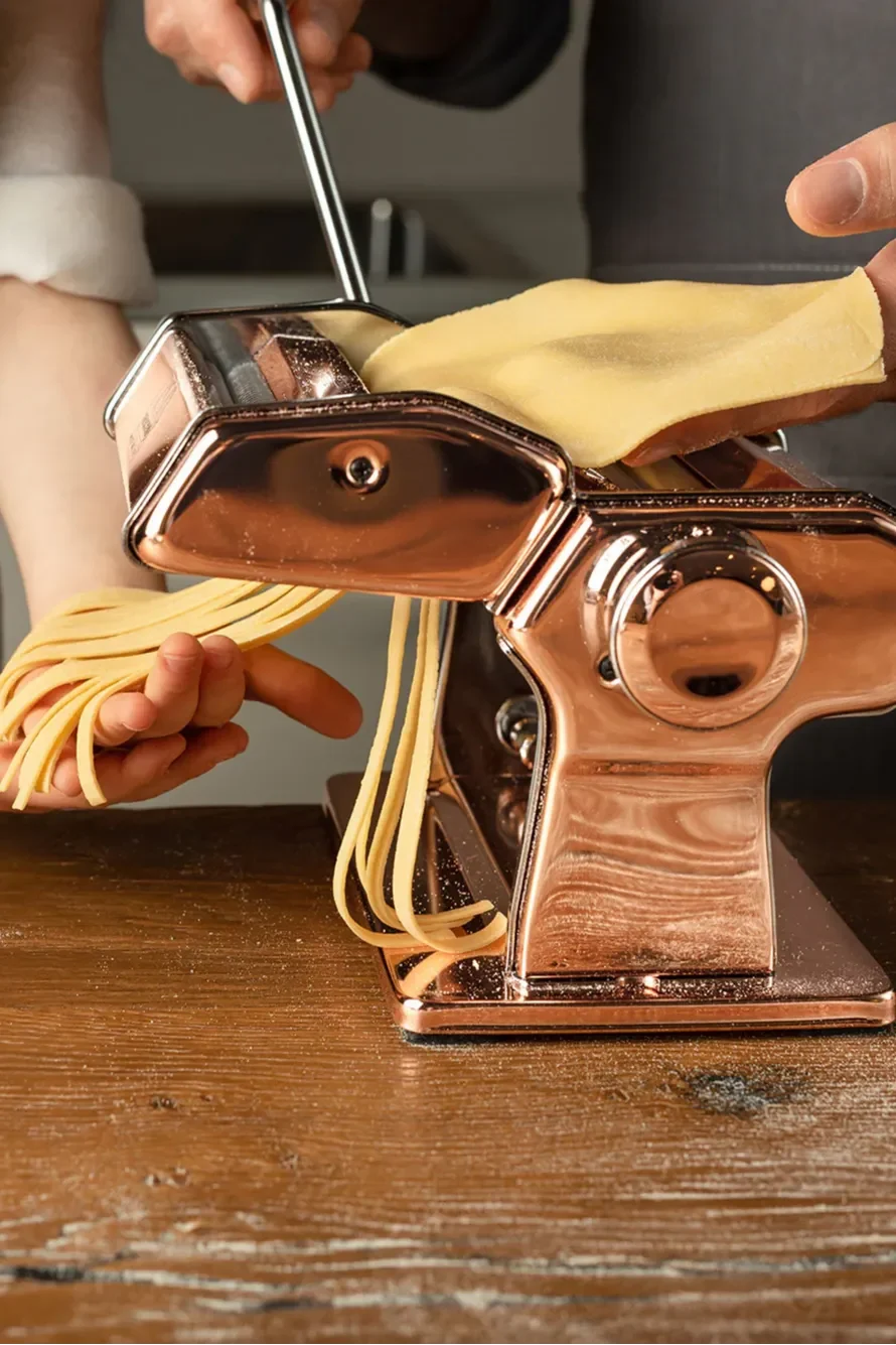 Hände bedienen eine kupferfarbene Nudelmaschine, führen Teig ein und produzieren frische Nudelstränge auf einem Holztisch.