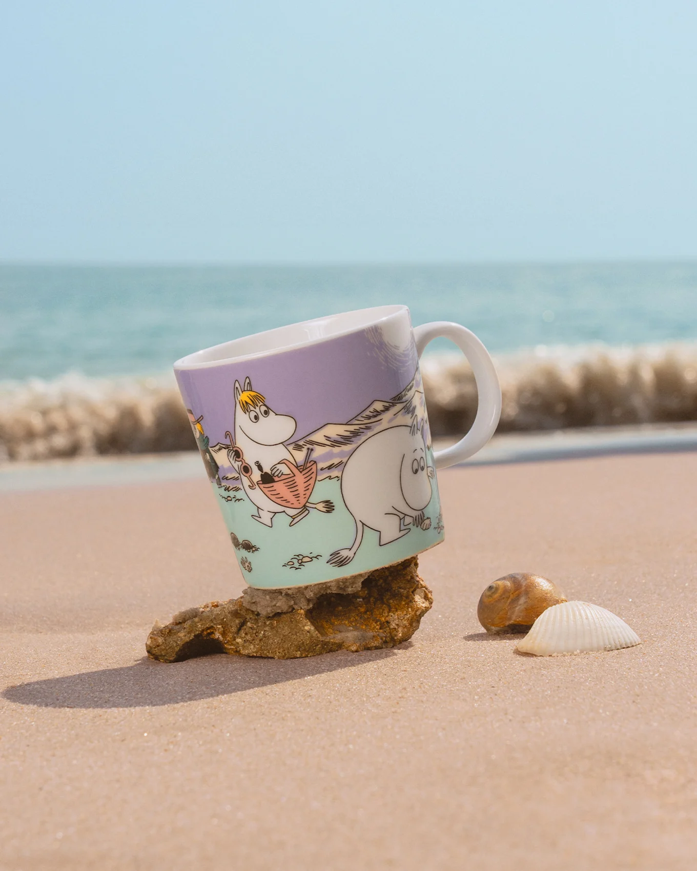 Mumin-Tasse am Strand, mit Snorkfräulein und Mumin, auf Stein gekippt, mit Muscheln und Meereswellen.