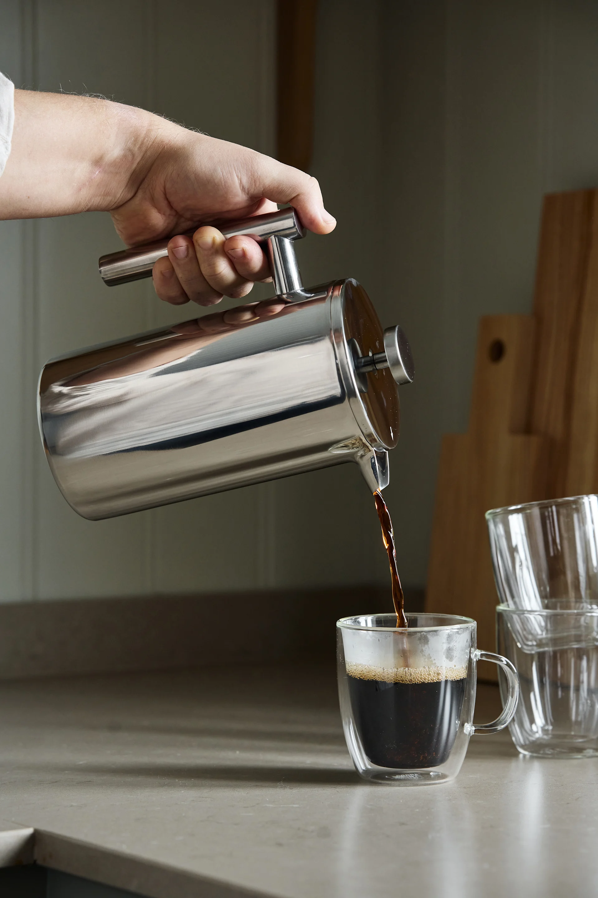 Hand gießt dunklen Kaffee aus einer Edelstahl-French Press in eine klare doppelwandige Glastasse auf einer Küchenarbeitsplatte.