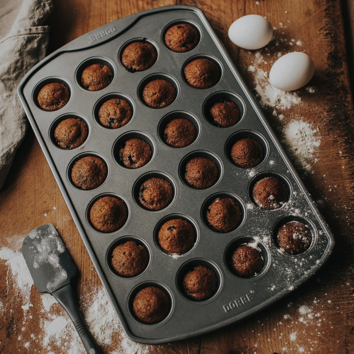 Eine Muffinform mit 24 gebackenen Mini-Muffins auf einer Holzoberfläche mit Mehl, zwei weißen Eiern und einem Spatel.
