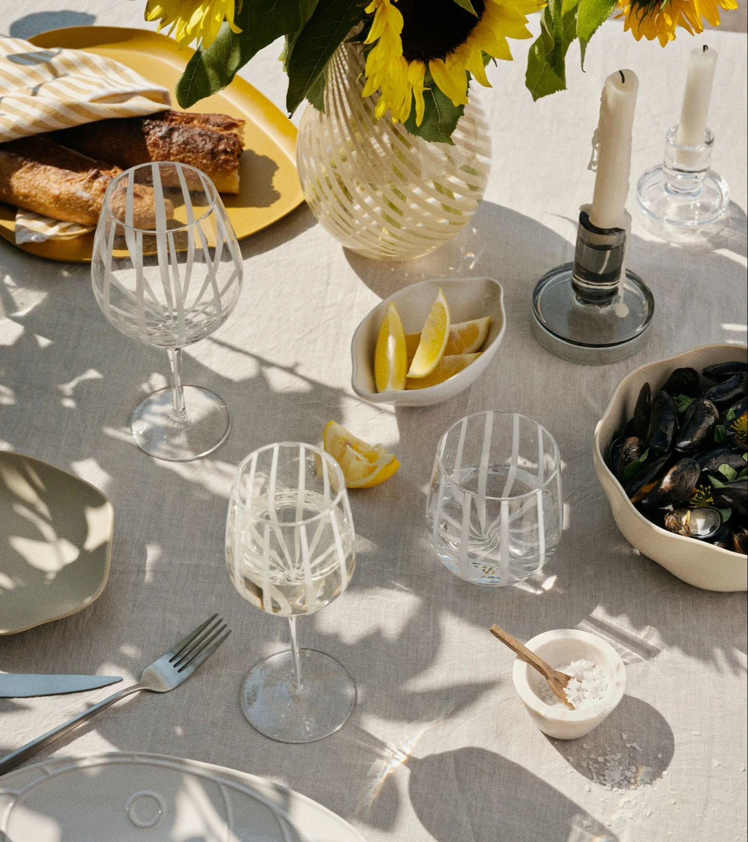 Eine sonnige Außentischdekoration mit Baguettes, Muscheln, Zitronen, Weingläsern, Sonnenblumen und Kerzen.