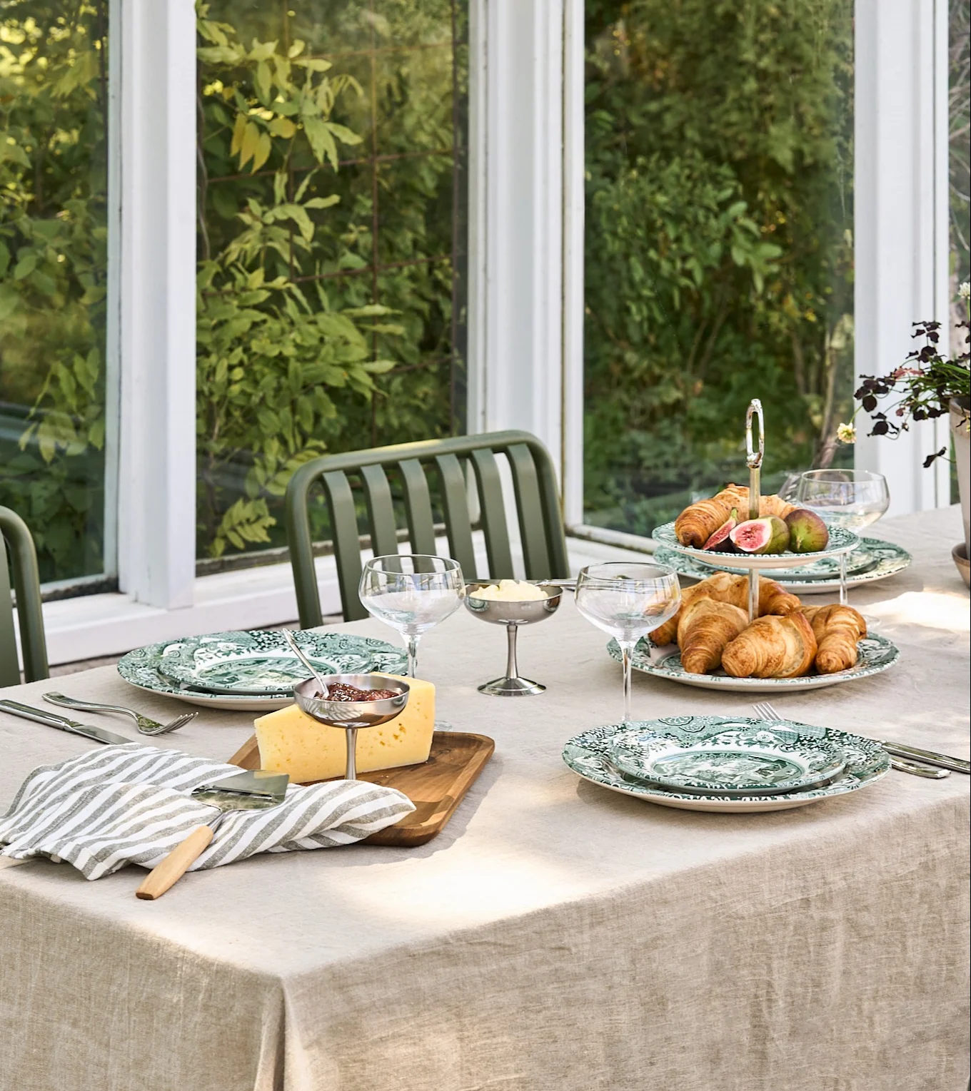 Ein Frühstückstisch mit grün gemusterten Tellern, Croissants, Feigen, Käse, Marmelade und Butter in einem hellen Wintergarten.