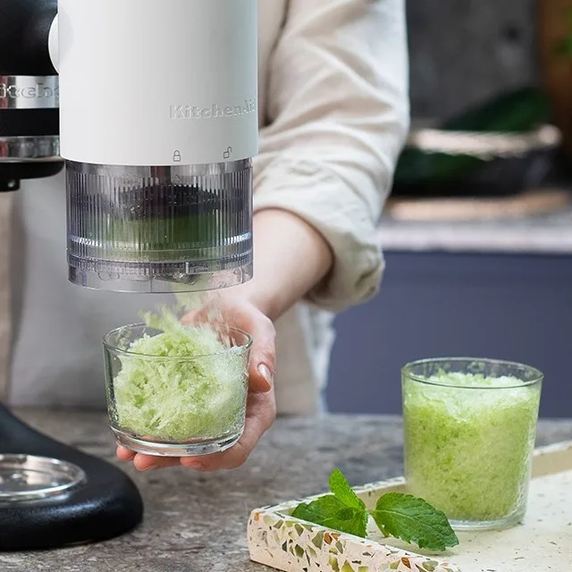 Eine Person verwendet ein KitchenAid-Zubehör, um eine grüne Substanz in ein Glas zu raspeln, mit einem weiteren Glas und Minzblättern daneben.