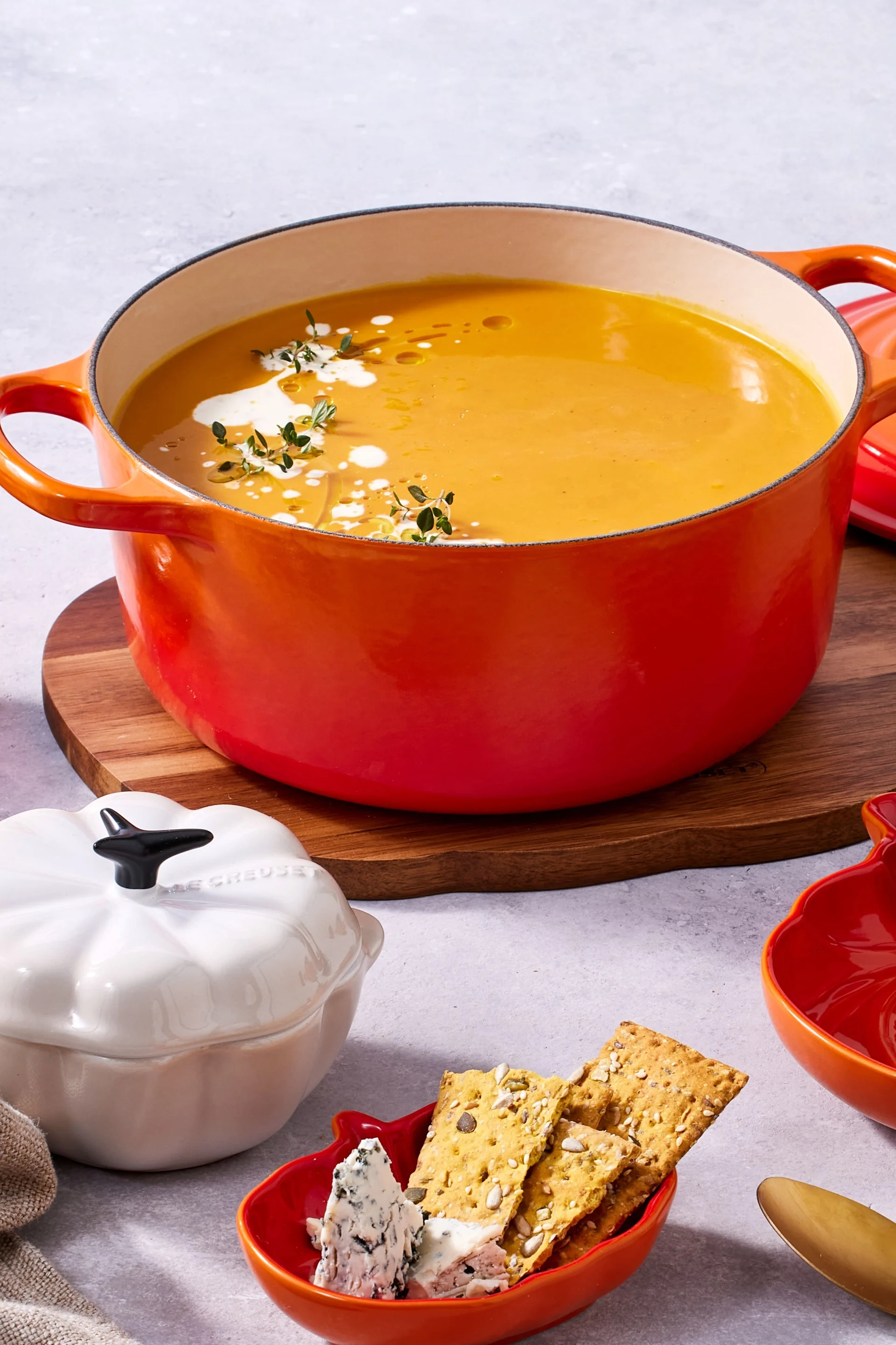 Oranger Topf mit cremiger Kürbissuppe, garniert mit Sahne und Thymian, dazu Cracker und Blauschimmelkäse in einer kürbisförmigen Schüssel.