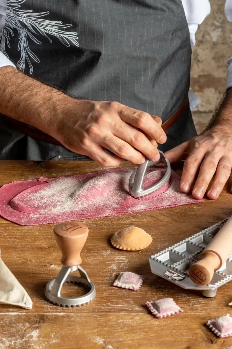 Hände verwenden einen Nudelschneider auf rosa Teig mit Mehl, machen Ravioli auf einem Holztisch mit Werkzeugen.