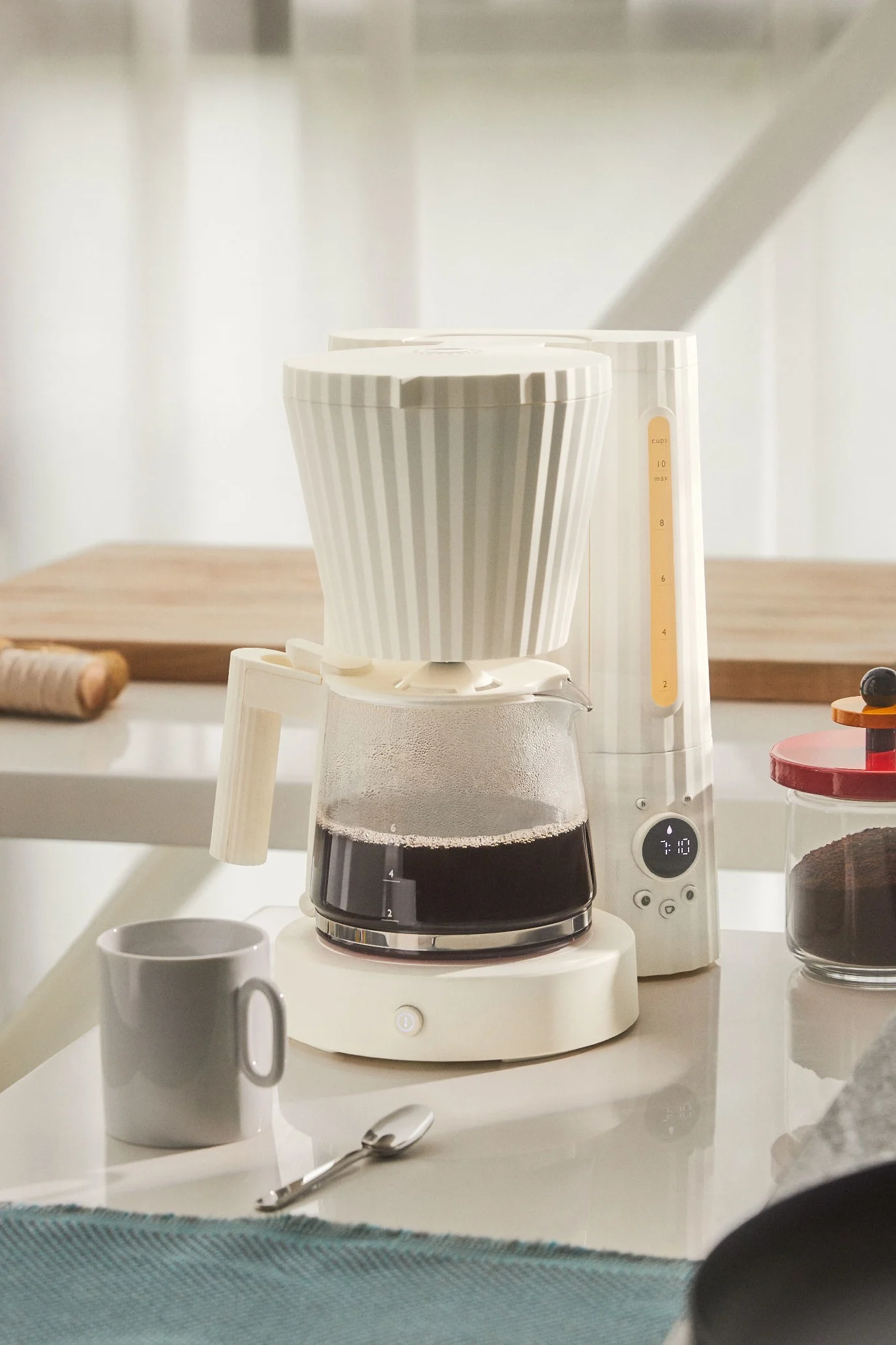 Eine hellbeige, gerippte Kaffeemaschine mit Glaskanne voll Kaffee steht auf einer weißen Arbeitsplatte neben einem grauen Becher.
