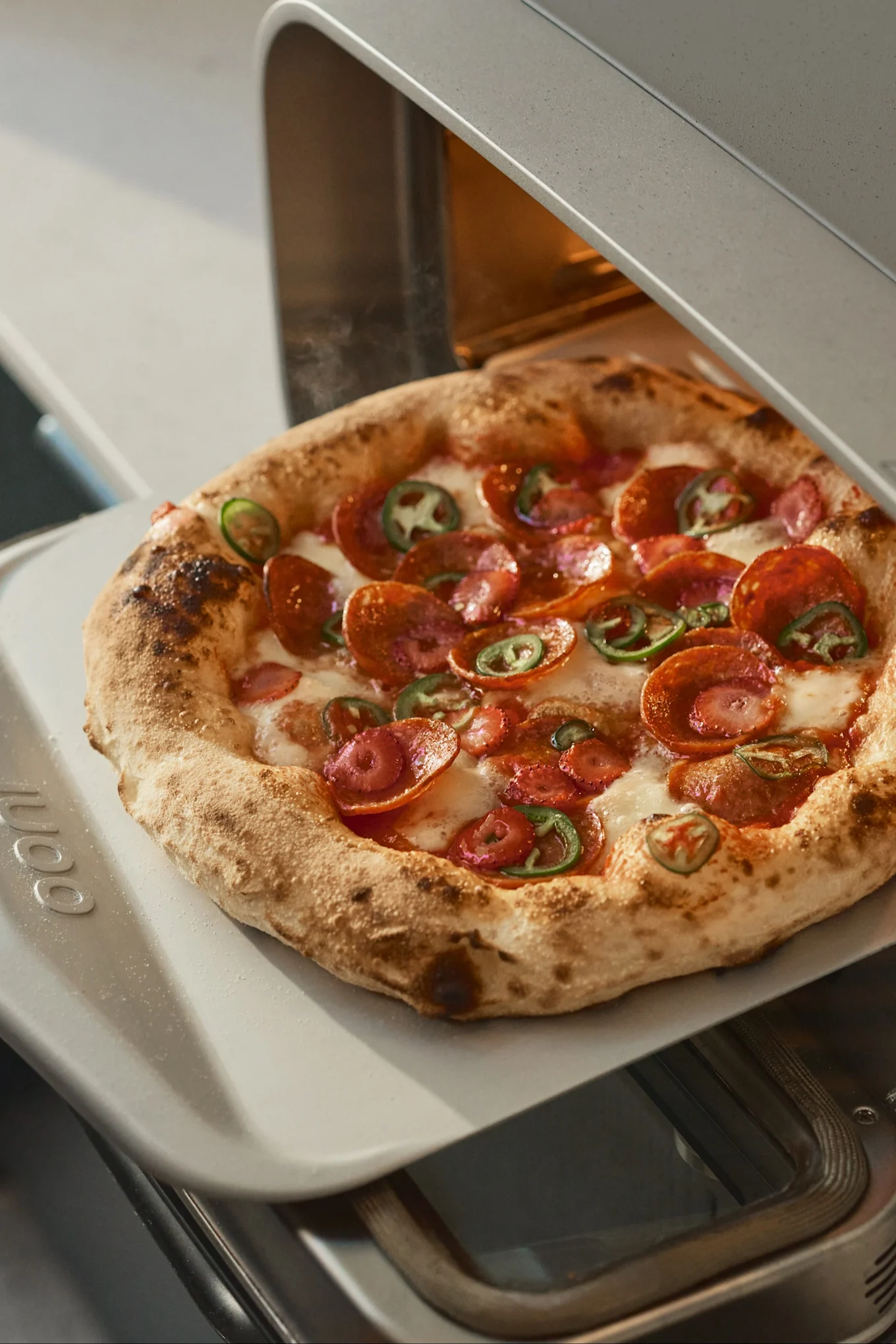 Eine Hand zieht eine Peperoni- und Jalapeño-Pizza mit einem Pizzaschieber aus einem Pizzaofen.