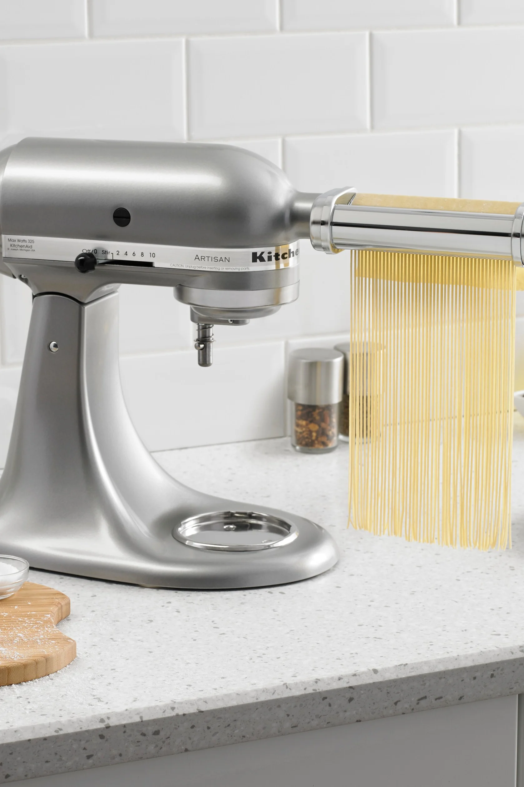 Eine silberne Küchenmaschine mit Nudelaufsatz, die frische Spaghetti auf eine helle, gesprenkelte Arbeitsplatte extrudiert.