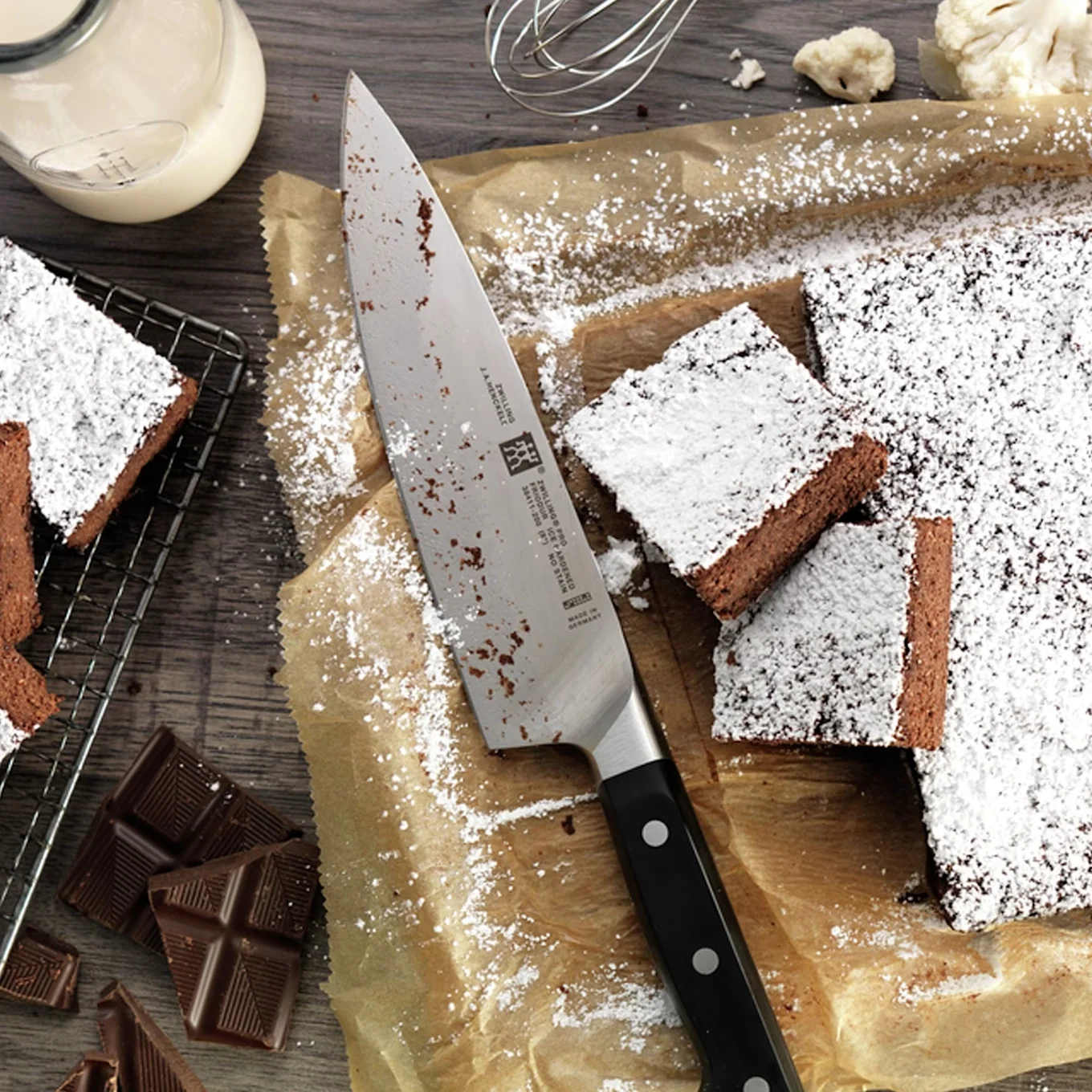 Frisch gebackene Schokoladenbrownies, mit Puderzucker bestäubt, neben Messer, Kakaopulver und Milch.