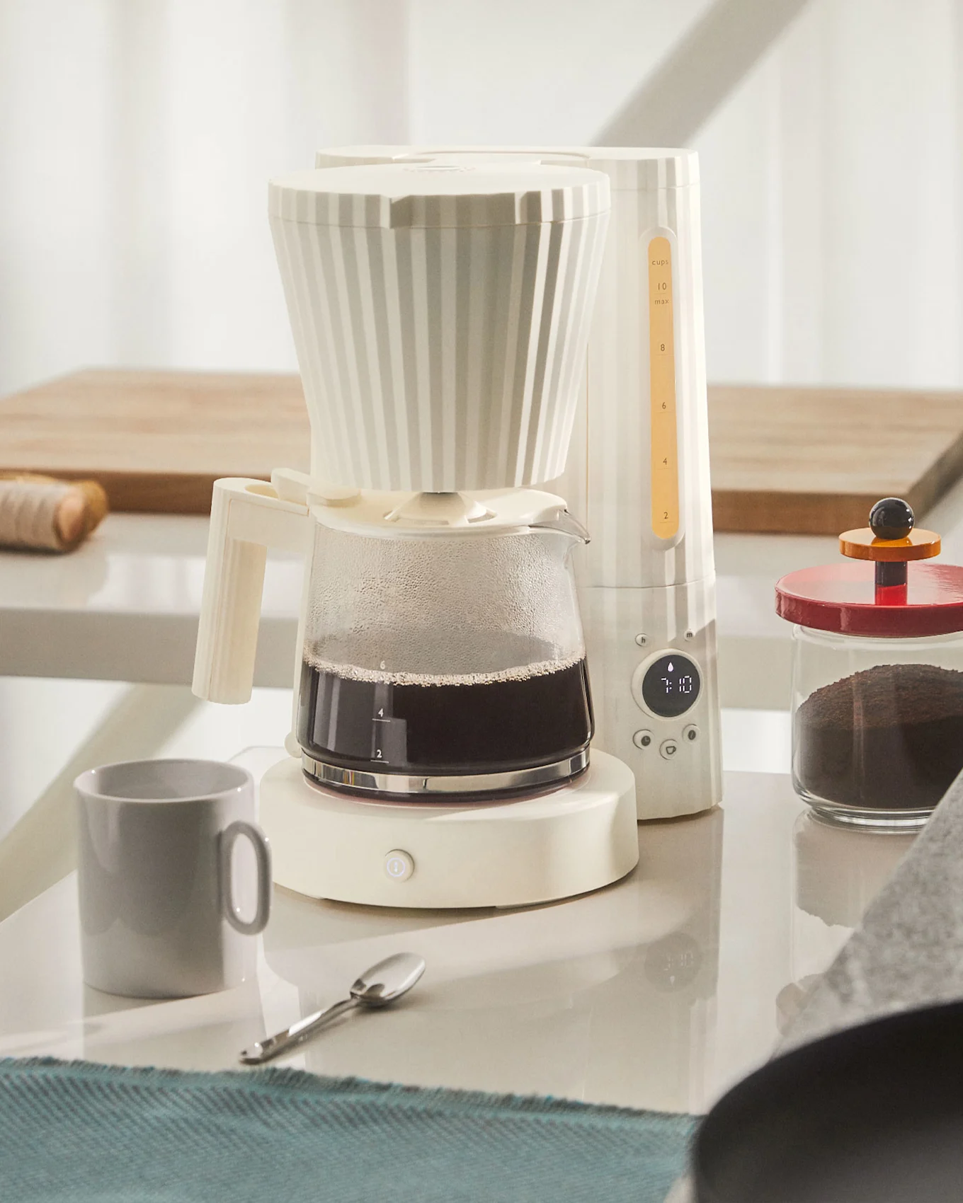 Cremefarbene Kaffeemaschine mit voller Glaskanne Kaffee, grauem Becher und Kaffeepulver auf weißer Theke.