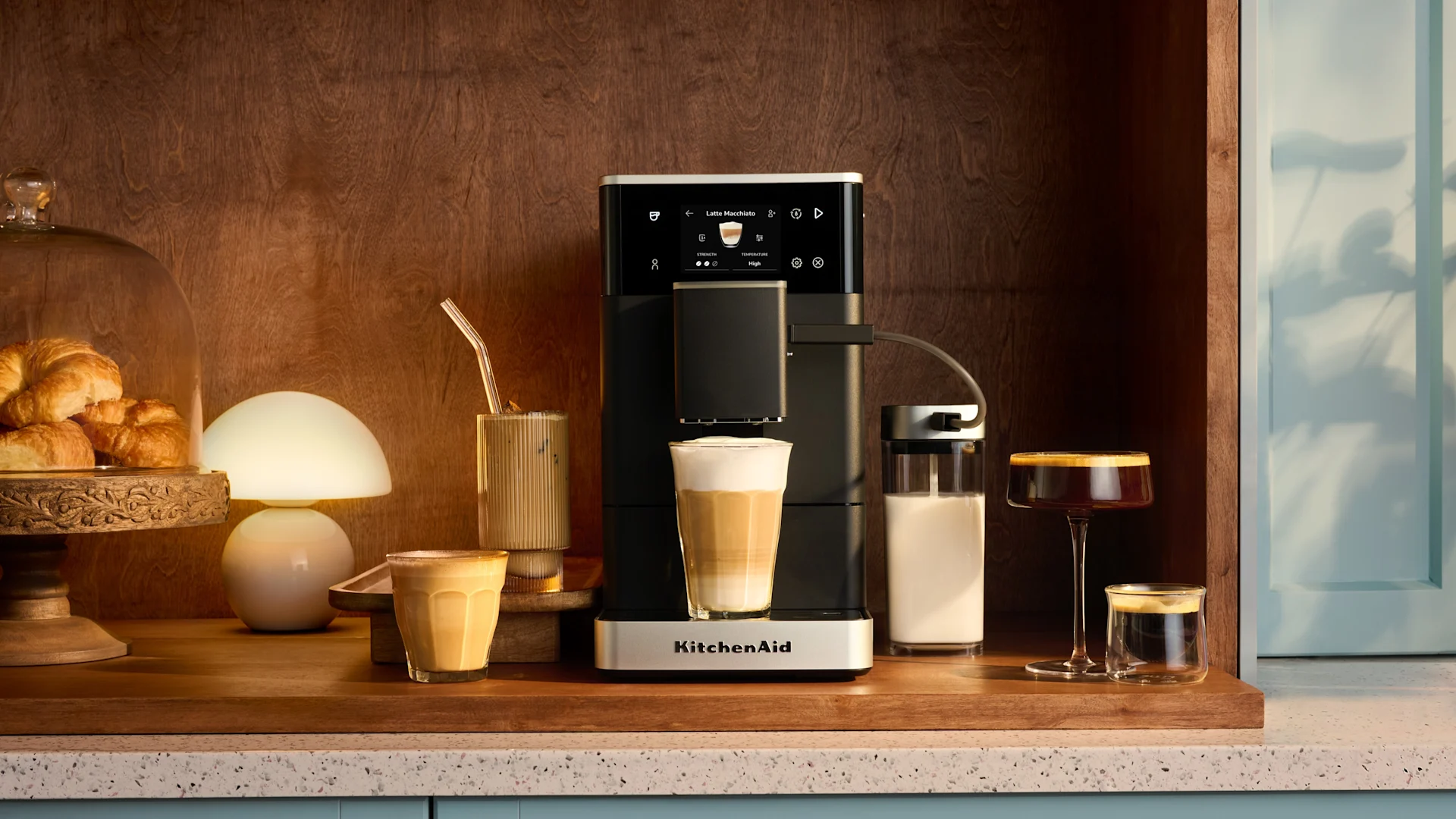 Eine Kaffeemaschine, die einen Latte Macchiato zubereitet, umgeben von Croissants und verschiedenen Kaffees auf einer Holztheke.