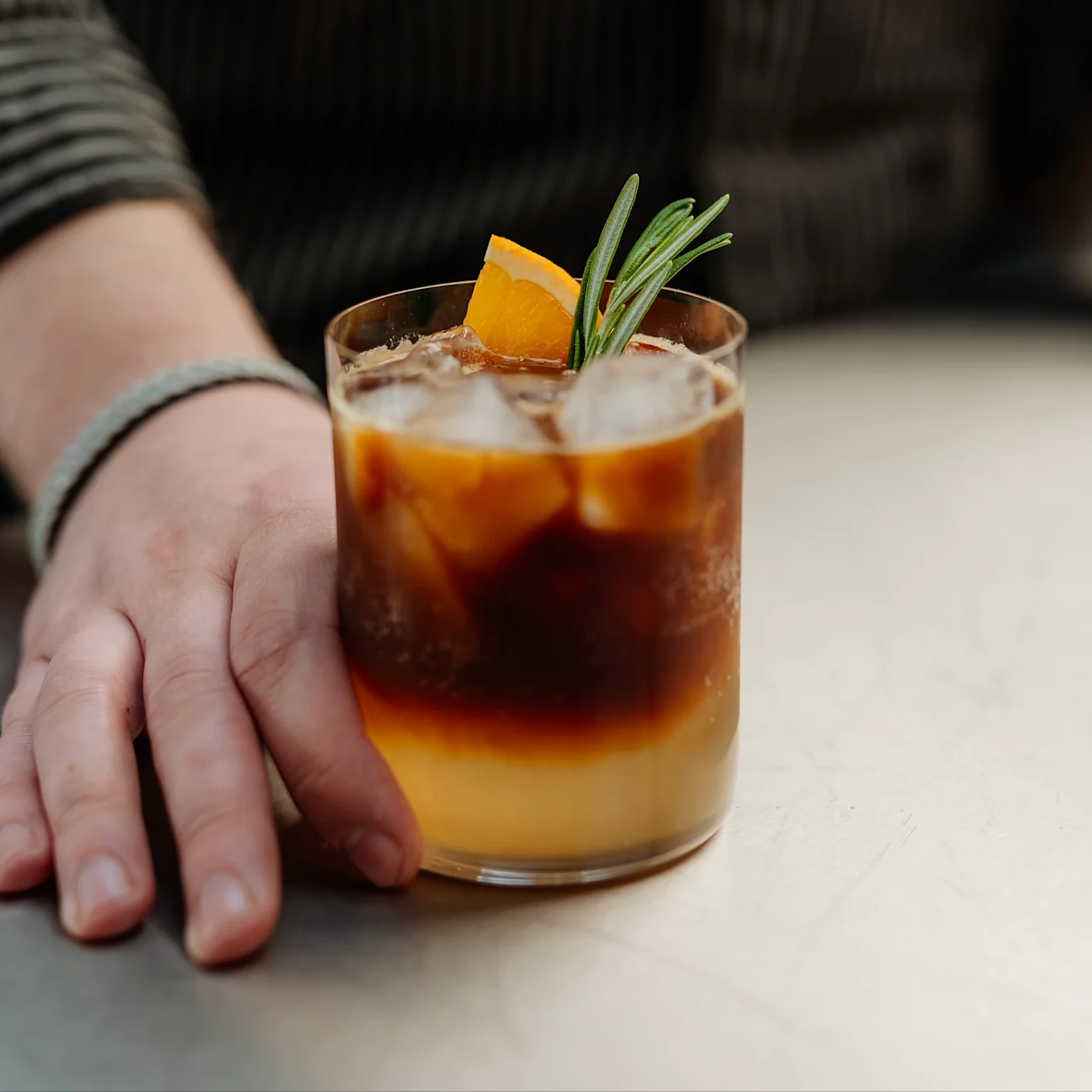 Eine Hand ruht neben einem geschichteten Eiskaffee mit Orangensaft, garniert mit einer Orangenscheibe und Rosmarin.