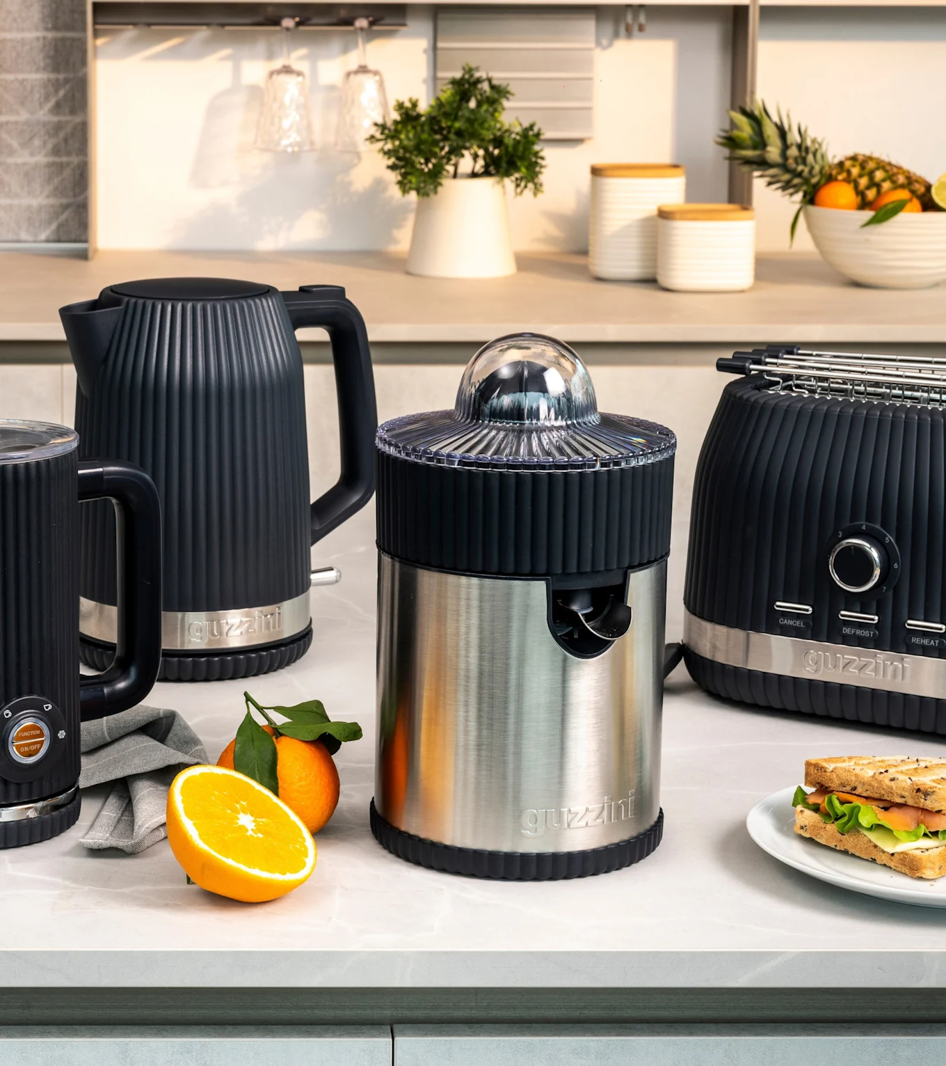 Schwarzer gerippter Wasserkocher, Milchaufschäumer, Edelstahl-Entsafter und Toaster auf Küchentheke mit Orangen und Sandwich.