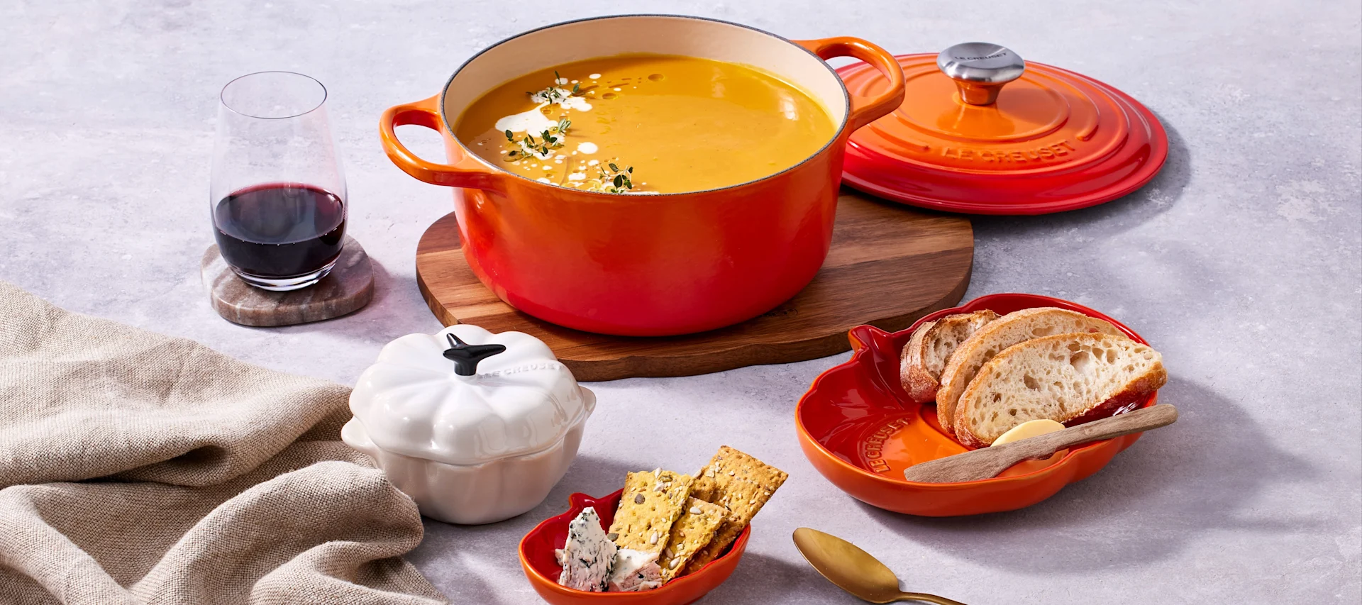 Ein oranger Topf mit cremiger gelber Suppe, garniert mit Kräutern und Sahne, steht auf einem Holzbrett neben dem Deckel, einem Glas Rotwein, Brot, Käse und Crackern.