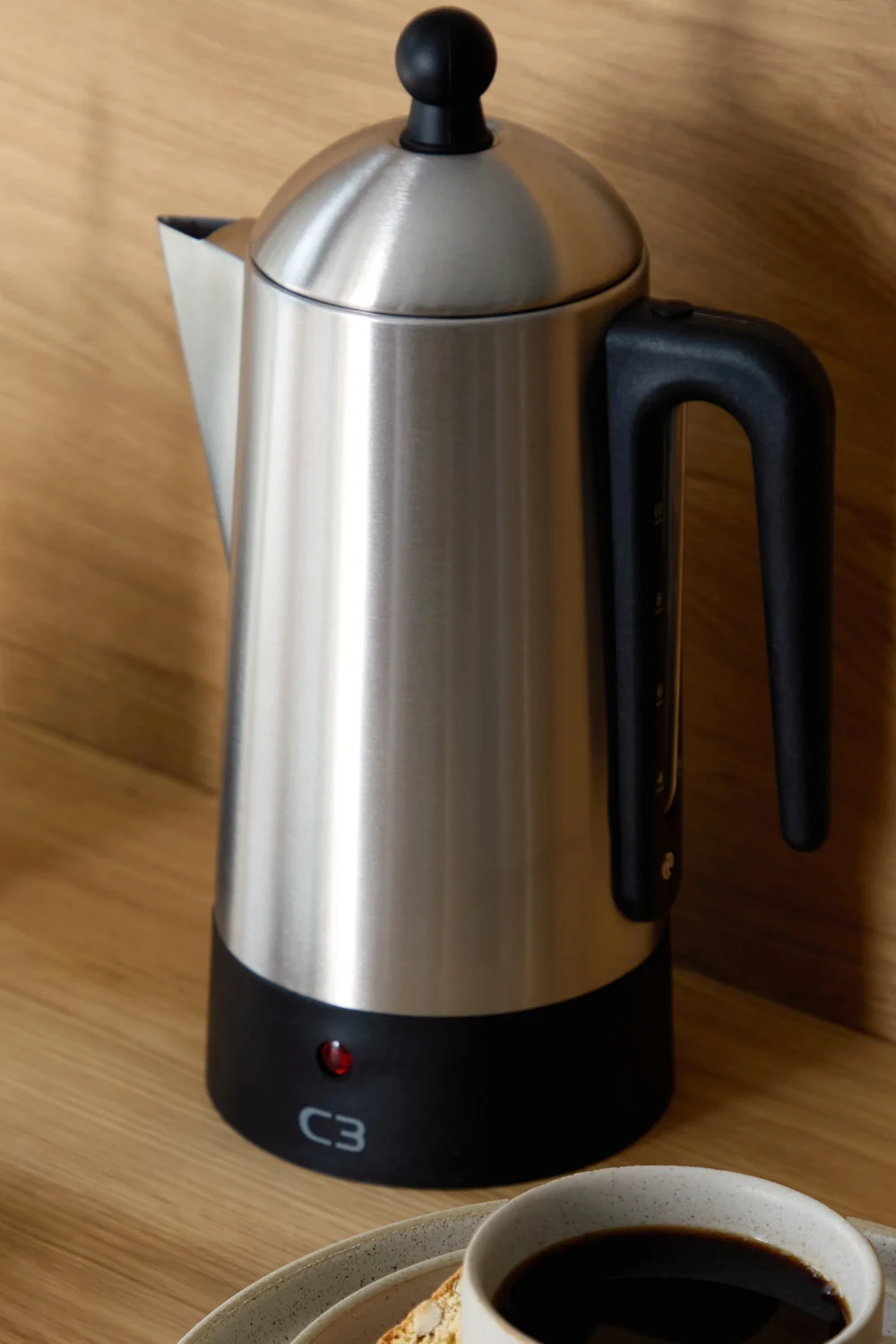 Eine Kaffeemaschine aus Edelstahl mit schwarzem Griff und Sockel steht auf einem Holztisch neben einer Tasse Kaffee und einem Keks.