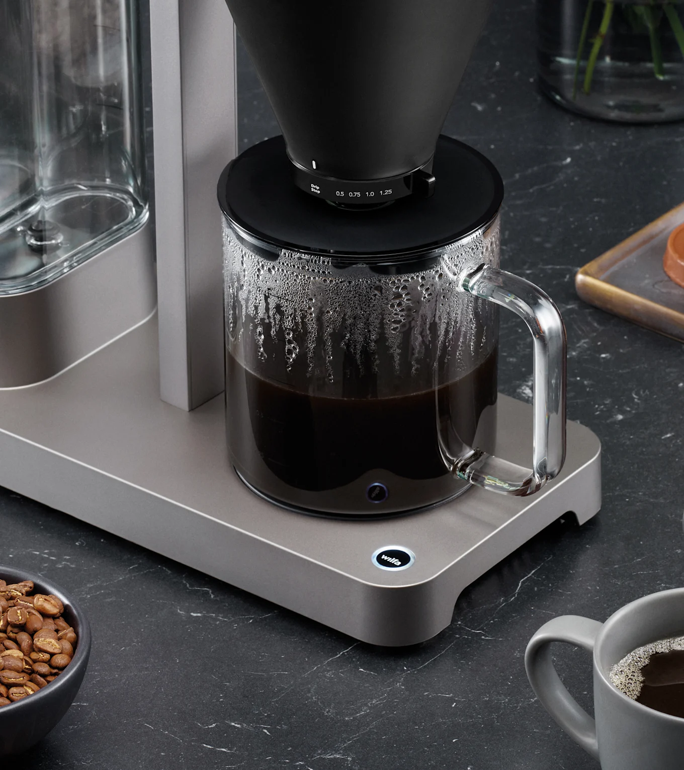 Eine moderne Kaffeemaschine mit schwarzem Filterkegel brüht Kaffee in eine Glaskaraffe auf dunkler Arbeitsplatte, mit Kaffeebohnen und Tasse.