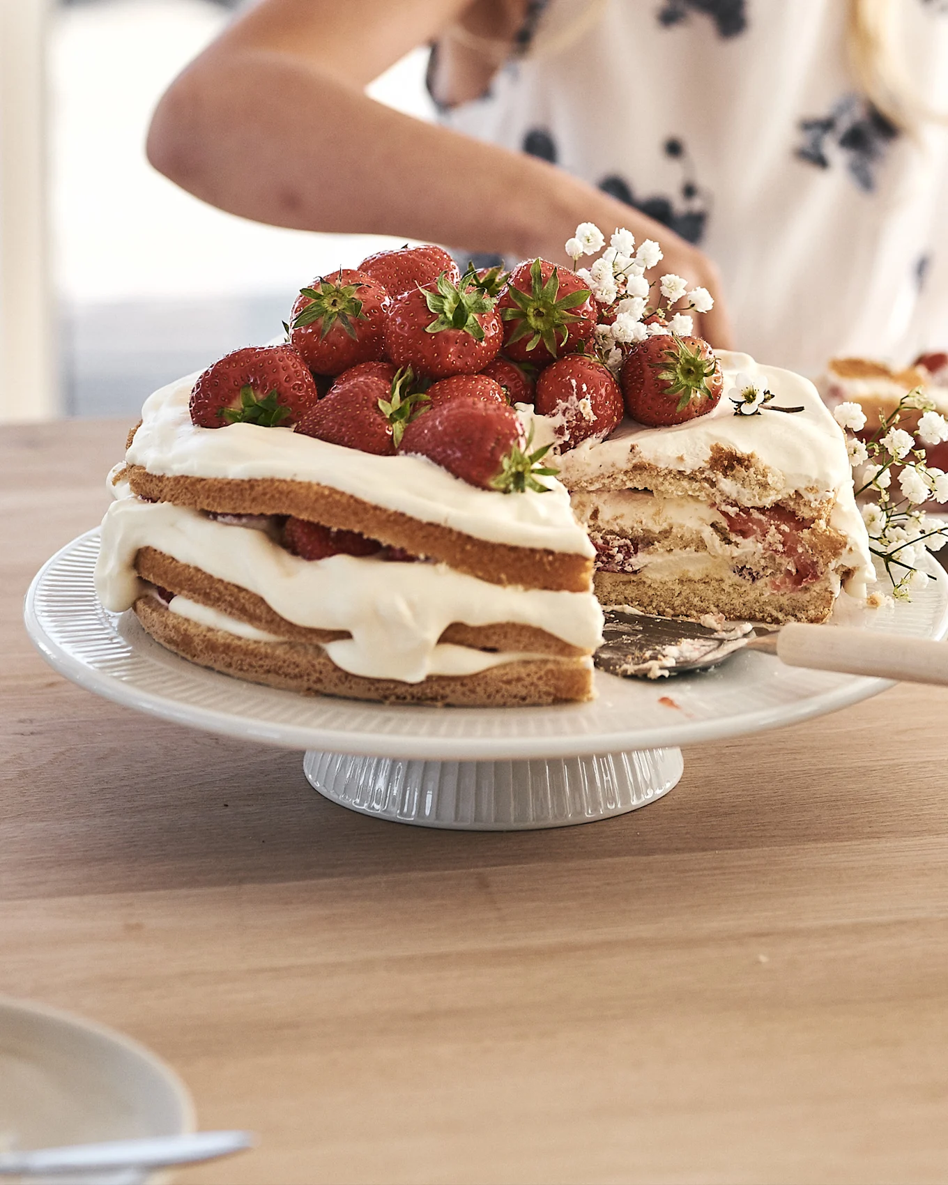 Eine Erdbeer-Sahnetorte mit frischen Erdbeeren auf einem weißen geriffelten Tortenständer. Ein Stück wurde herausgeschnitten.