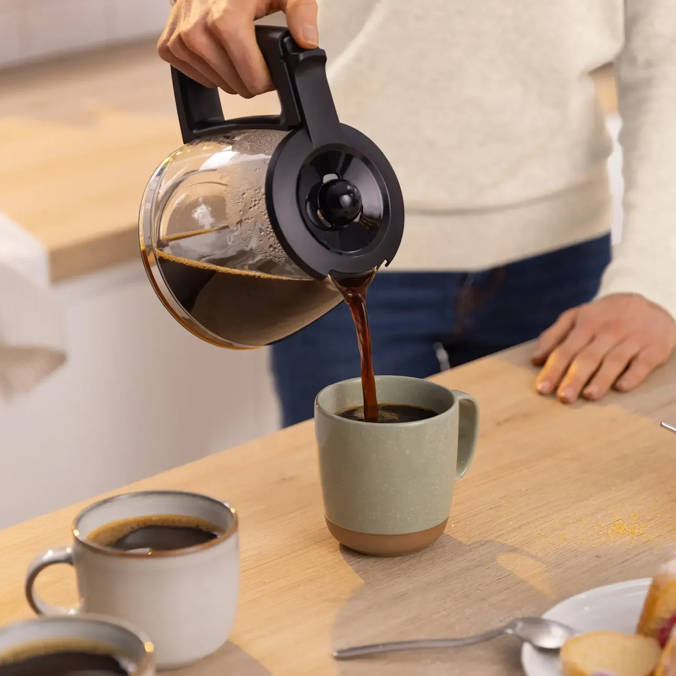 Eine Person gießt heißen Kaffee aus einer Glaskanne in eine hellgrüne Tasse auf einer Holztheke. Eine weitere Tasse ist im Vordergrund.