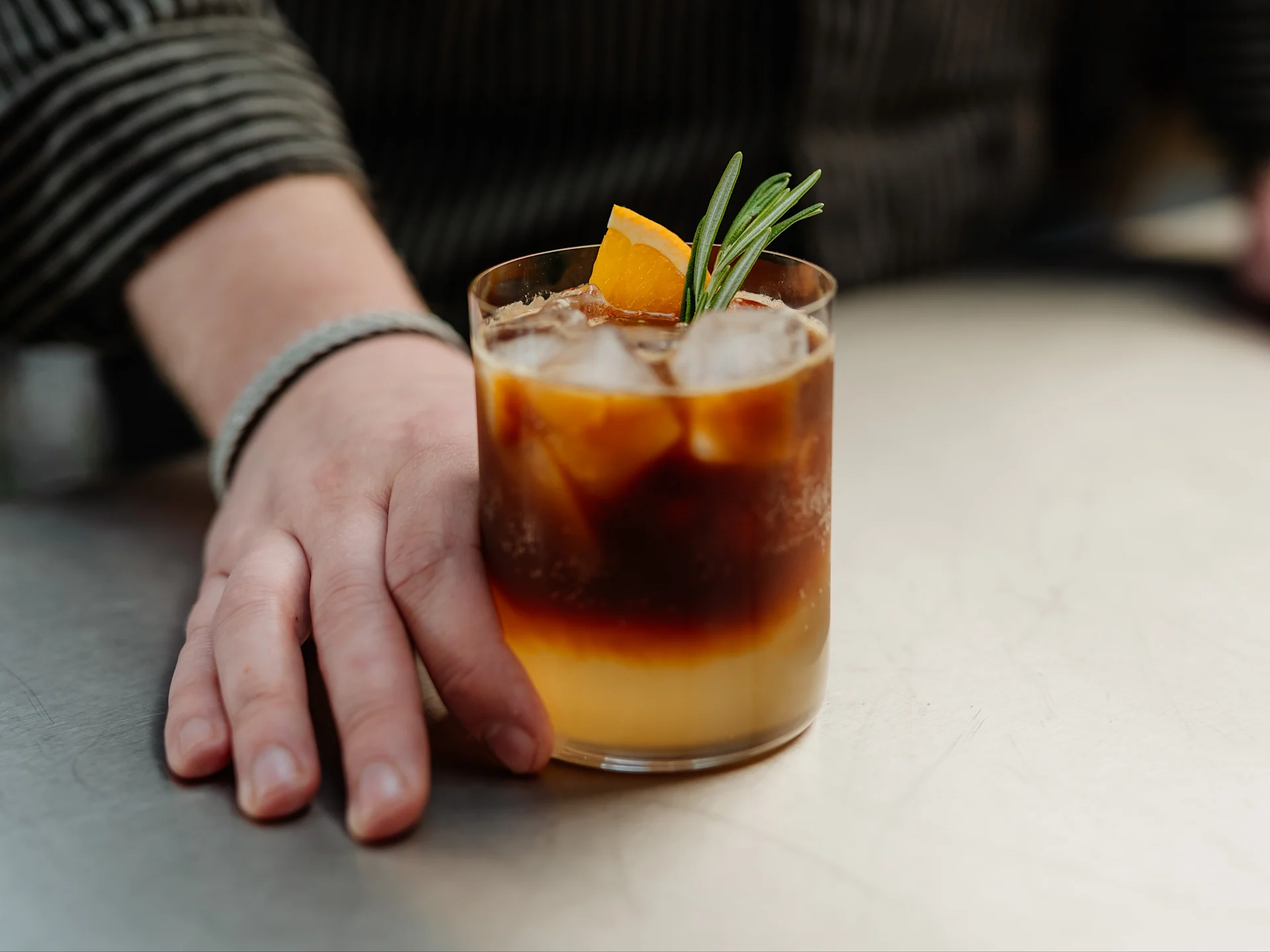 Eine Hand neben einem geschichteten Eiskaffee, garniert mit Orangenscheibe und Rosmarin, auf einer Metalloberfläche.