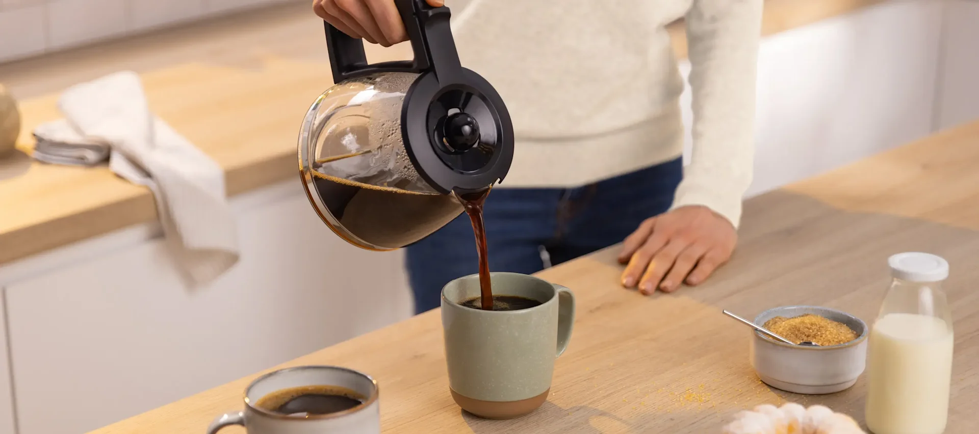 Person gießt dunklen Kaffee aus einer Glaskaraffe in einen hellgrünen Becher auf einer Holztheke mit Milch und Zucker.