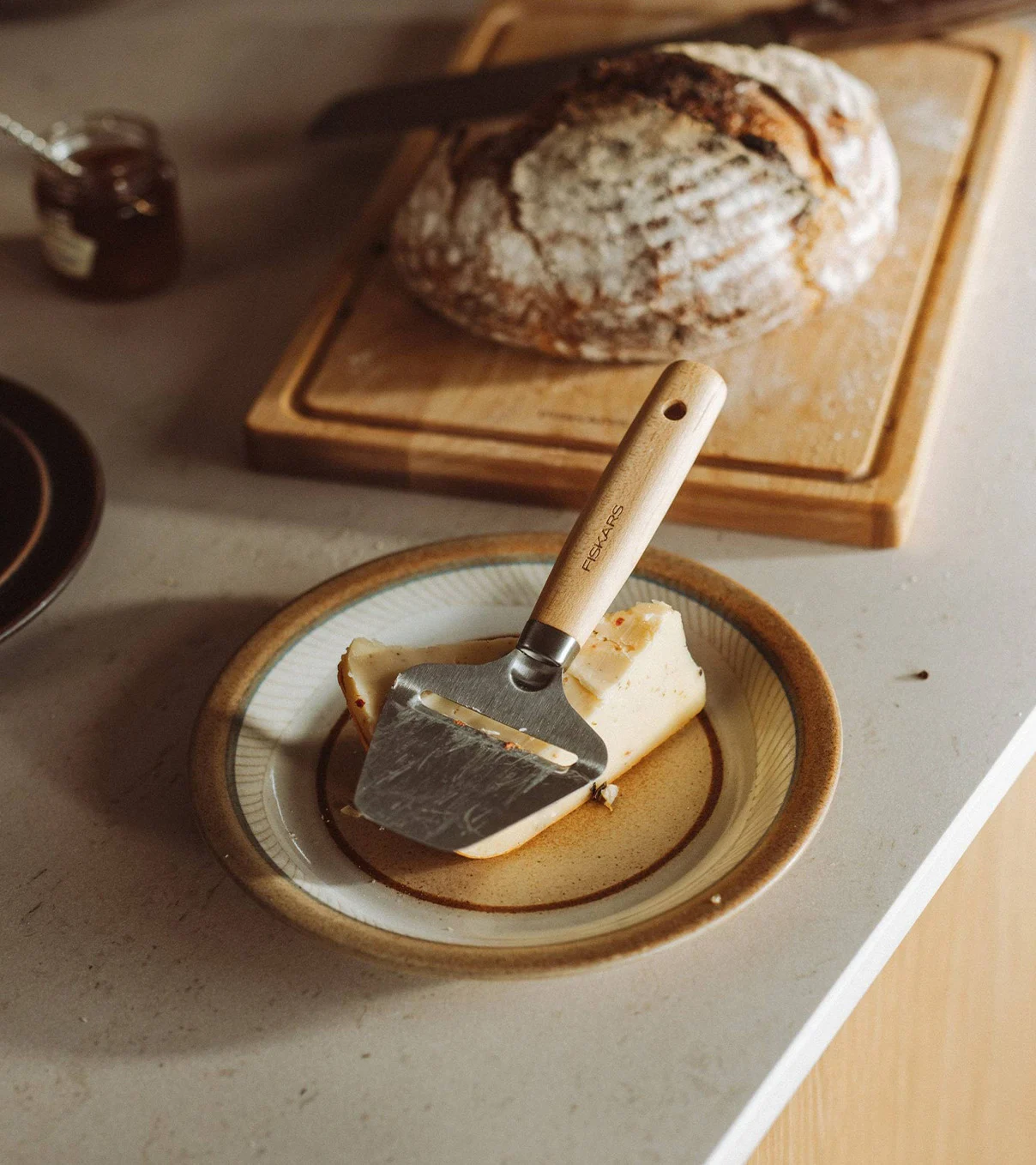 Ein Käsehobel mit Holzgriff liegt auf einem Stück Käse auf einem Teller, mit einem Laib Brot im Hintergrund.