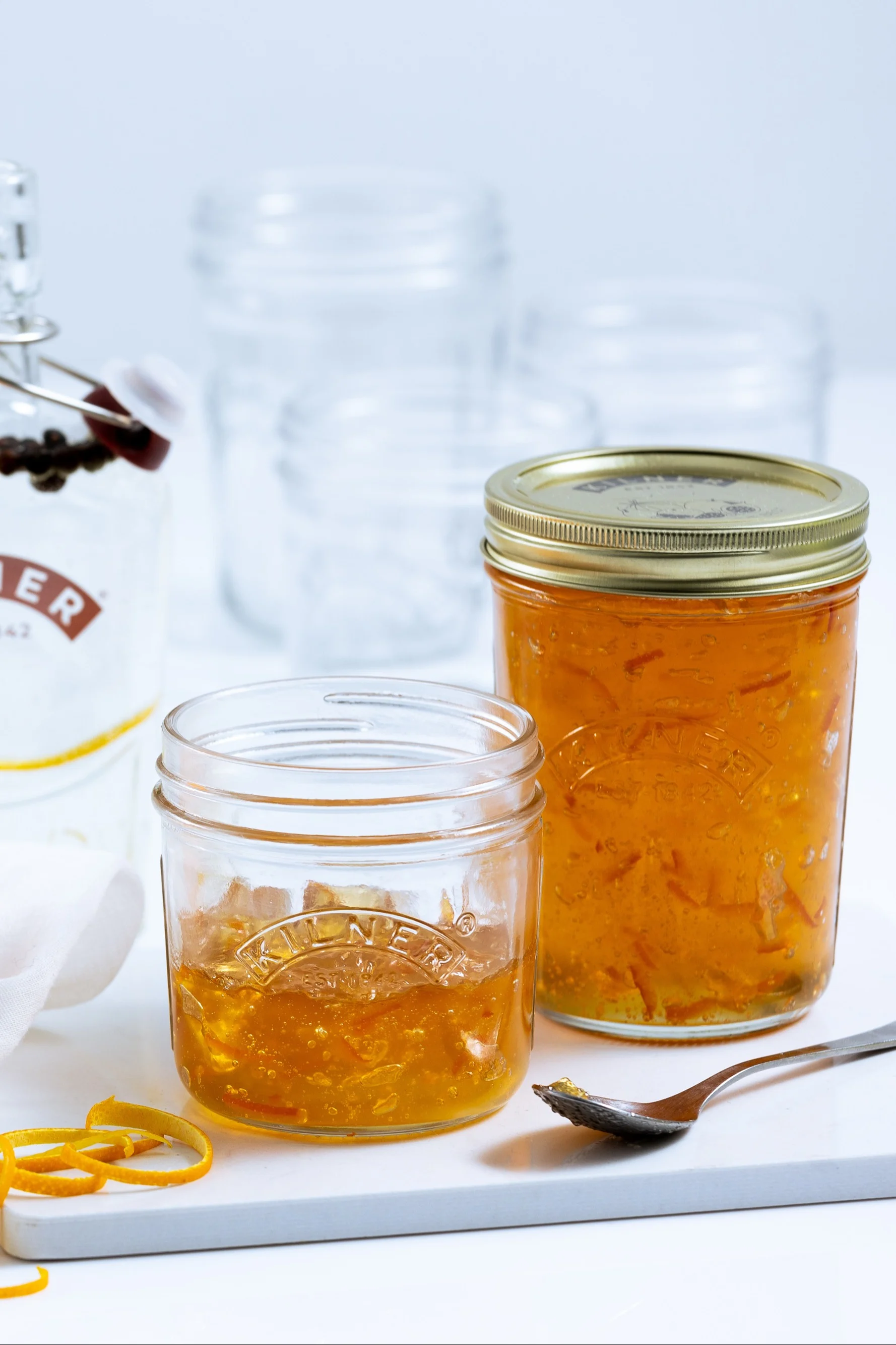 Zwei Gläser hausgemachte Orangenmarmelade auf weißer Oberfläche, eines offen mit einem Löffel, umgeben von Orangenschalen und leeren Gläsern.