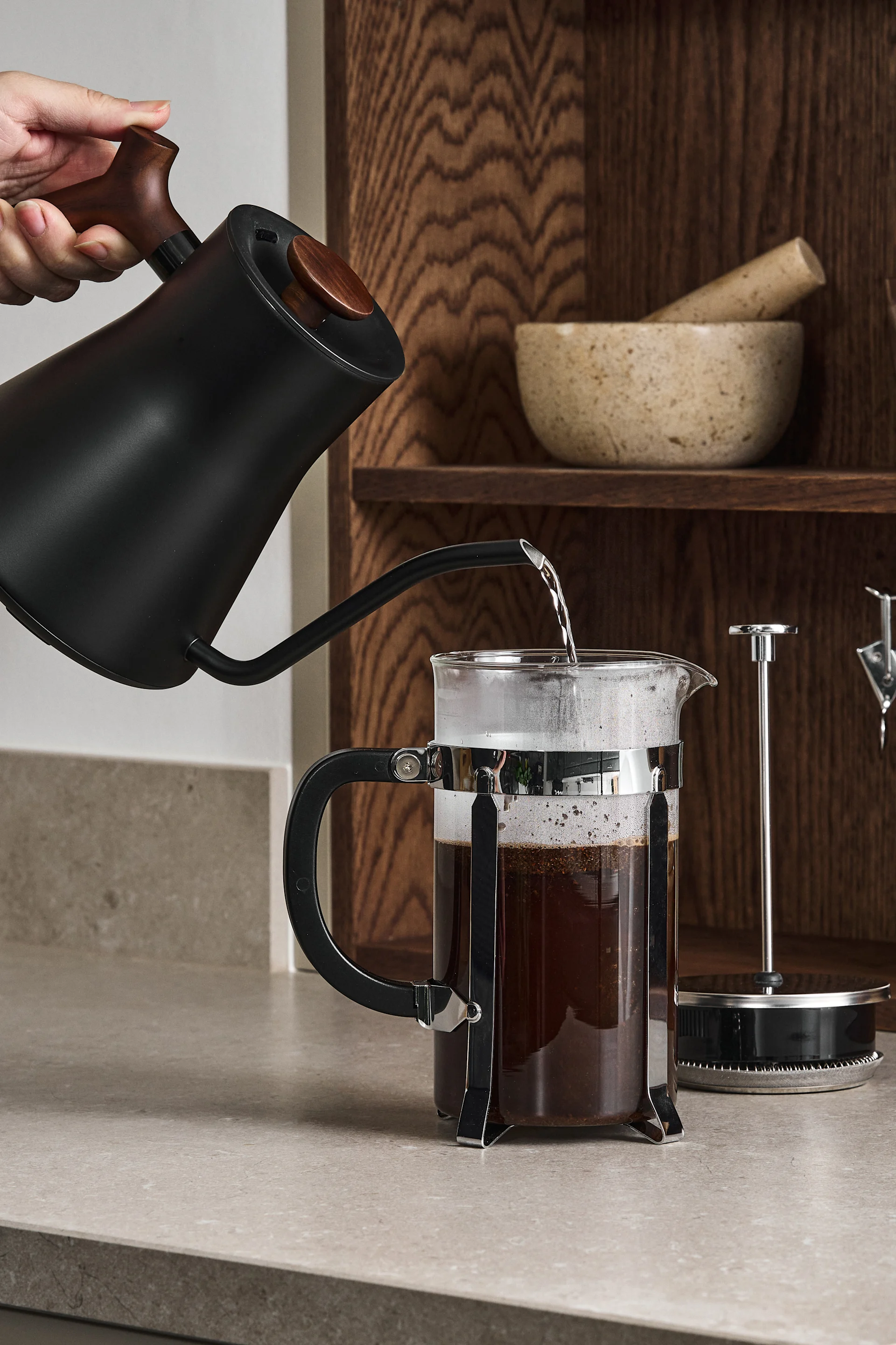 Eine Person gießt heißes Wasser aus einer schwarzen Schwanenhalskanne in eine French Press mit Kaffee auf einer Arbeitsplatte.