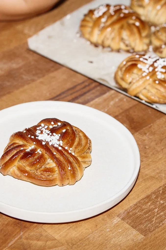 Eine Hand hält einen Teller mit einer einzelnen geflochtenen Brötchen, bestreut mit Hagelzucker. Ein Blech mit vielen ähnlichen Brötchen liegt im Hintergrund auf einem Holztisch.