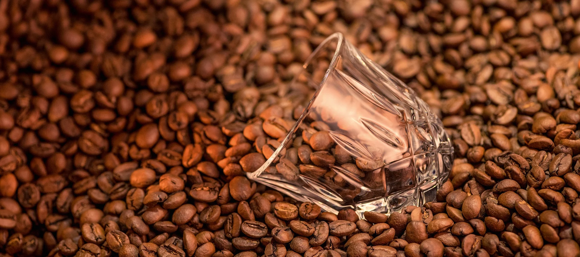 Kaffeebohnen mit einem umgekippten, leeren Glas in der Mitte, das das Licht der Kaffeebohnen reflektiert.