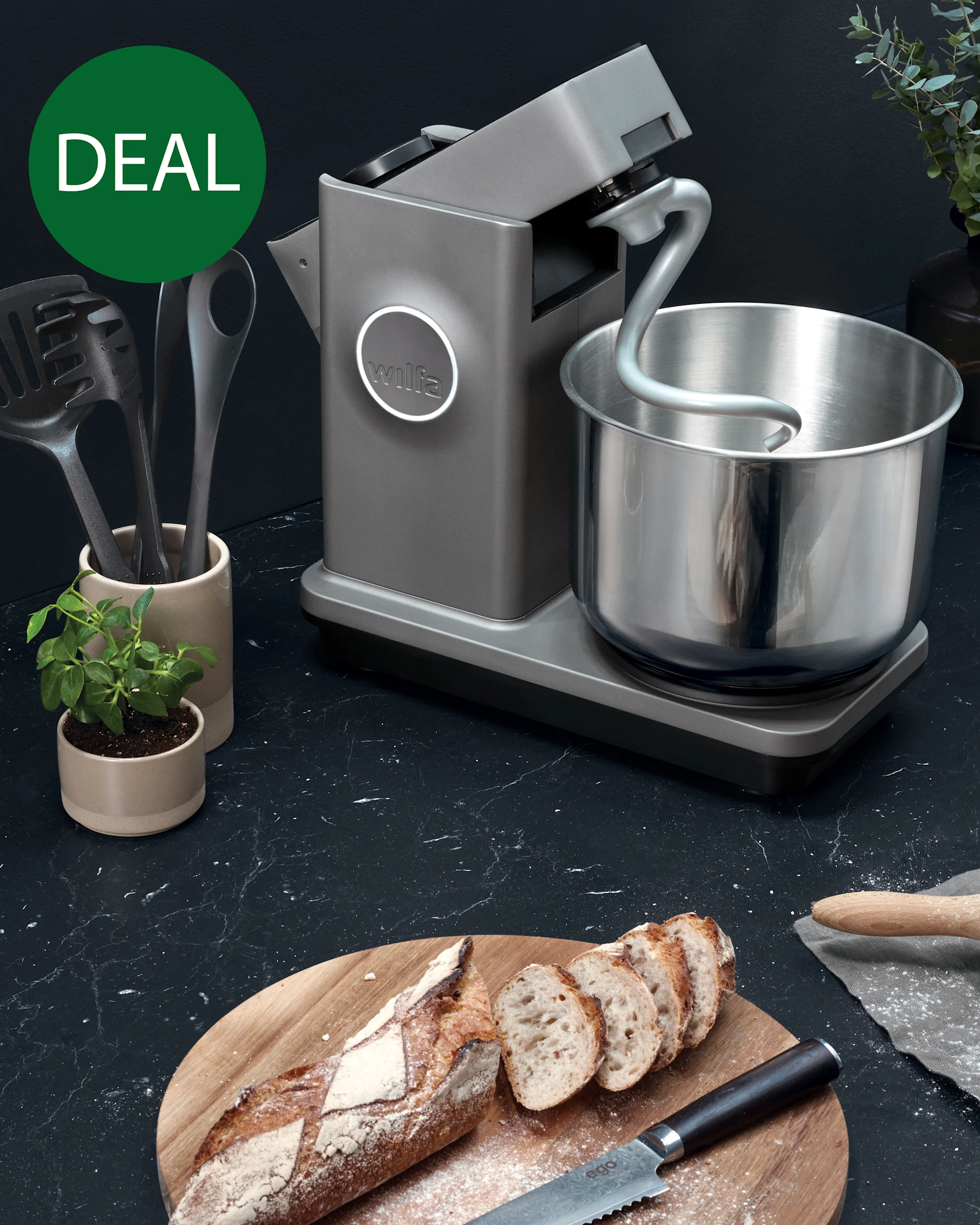 Ein grauer Küchenmaschine mit Edelstahlschüssel und Knethaken auf einer Küchentheke, neben geschnittenem Brot auf einem Brett. Ein grünes 'DEAL'-Etikett ist sichtbar.