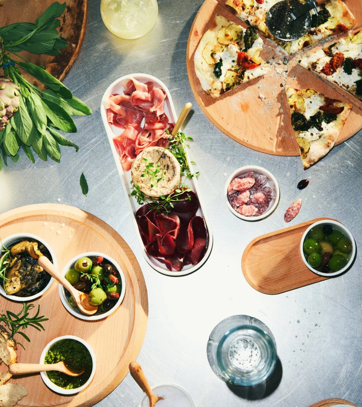 Blick von oben auf eine Essensauslage mit geschnittener Pizza, Wurstplatte, Oliven, Pesto und Tapenade auf Holzbrettern.