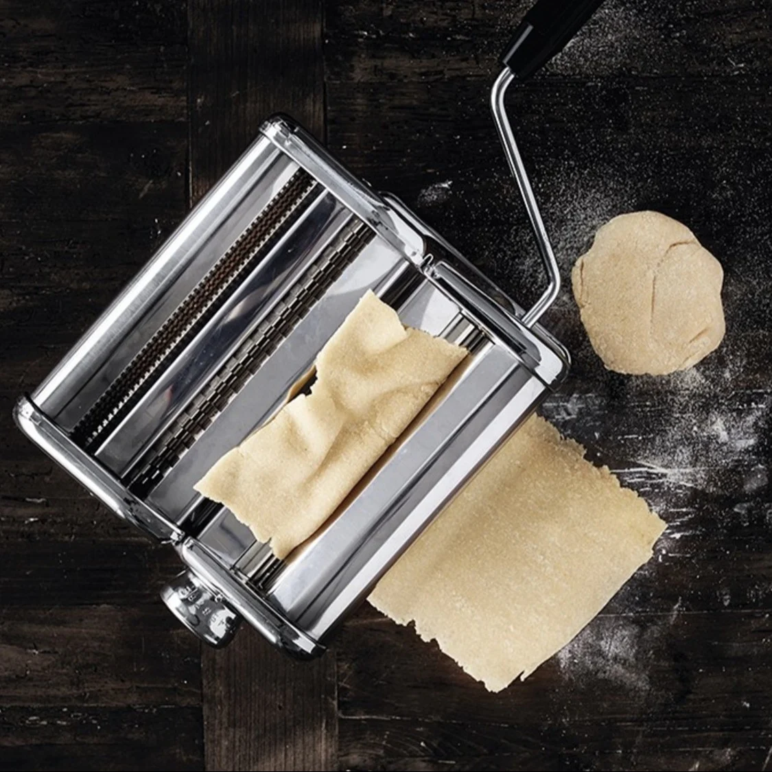 Eine silberne Nudelmaschine flacht Teig auf einem dunklen Holztisch mit einer Teigkugel und Mehl ab.