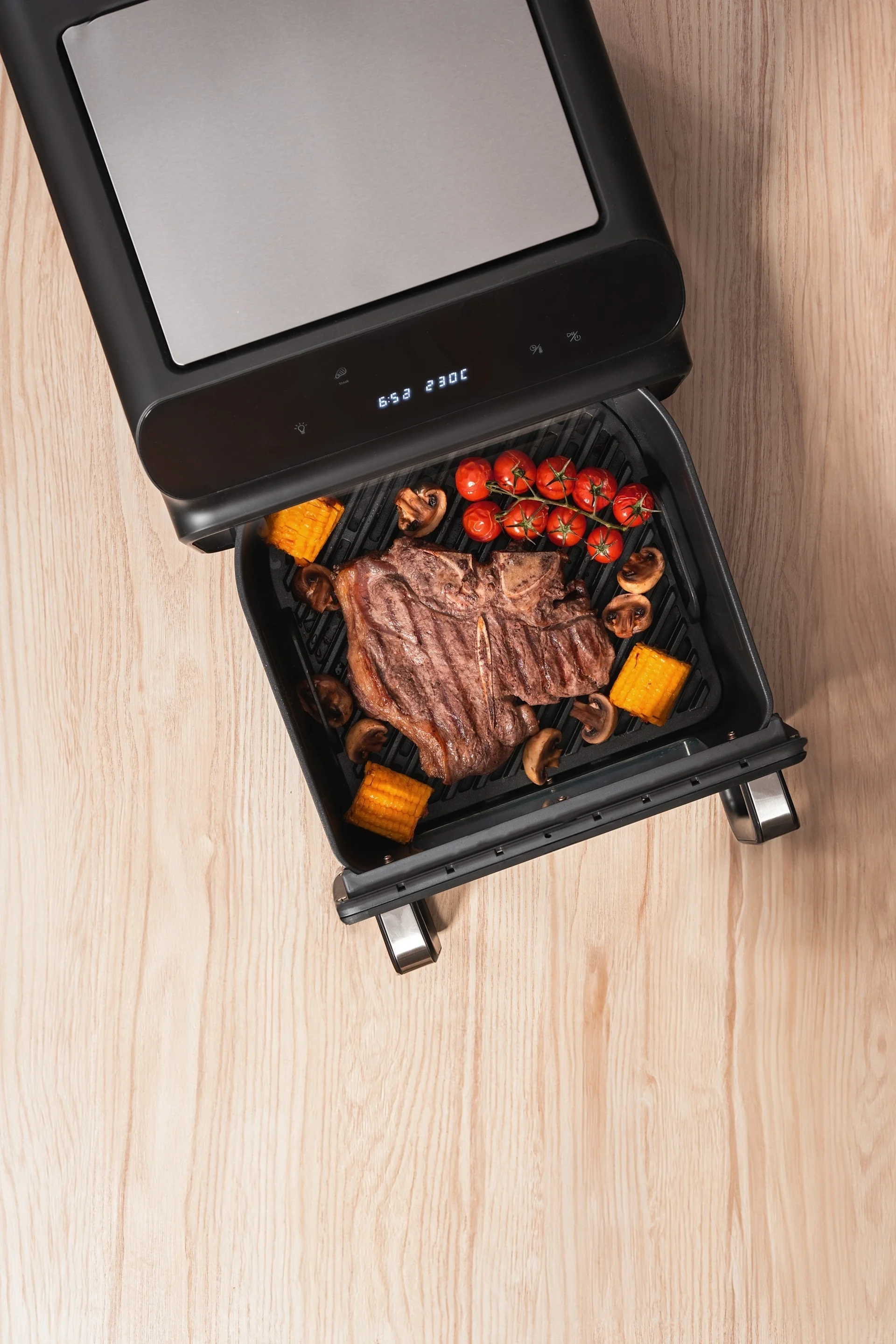 Ein T-Bone-Steak, Kirschtomaten, Pilze und Maiskolben garen in einem offenen Elektrogrill, der 230C anzeigt.