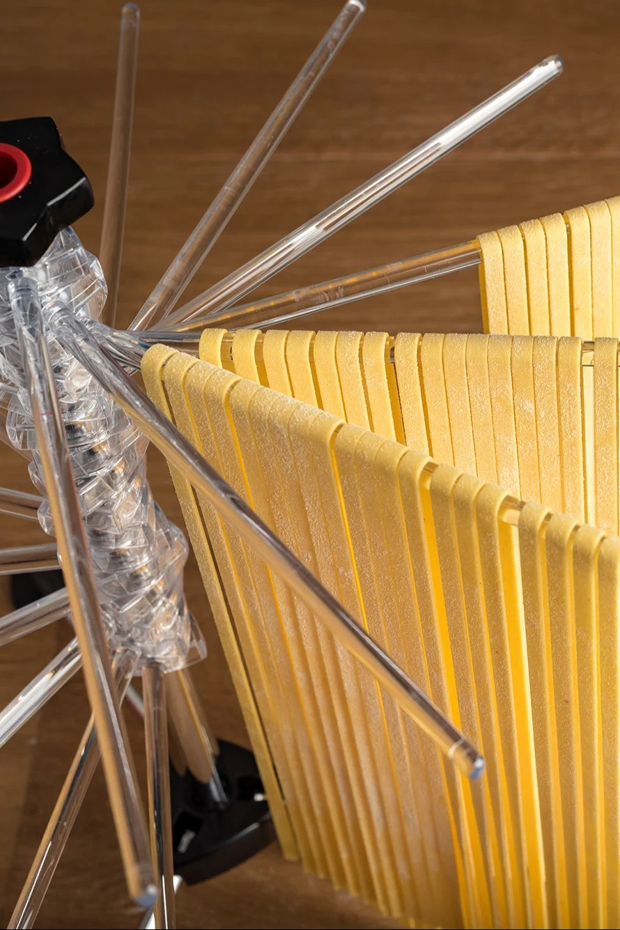 Frische gelbe Pasta trocknet auf einem transparenten Pastaständer mit mehreren Armen vor einem Holzhintergrund.