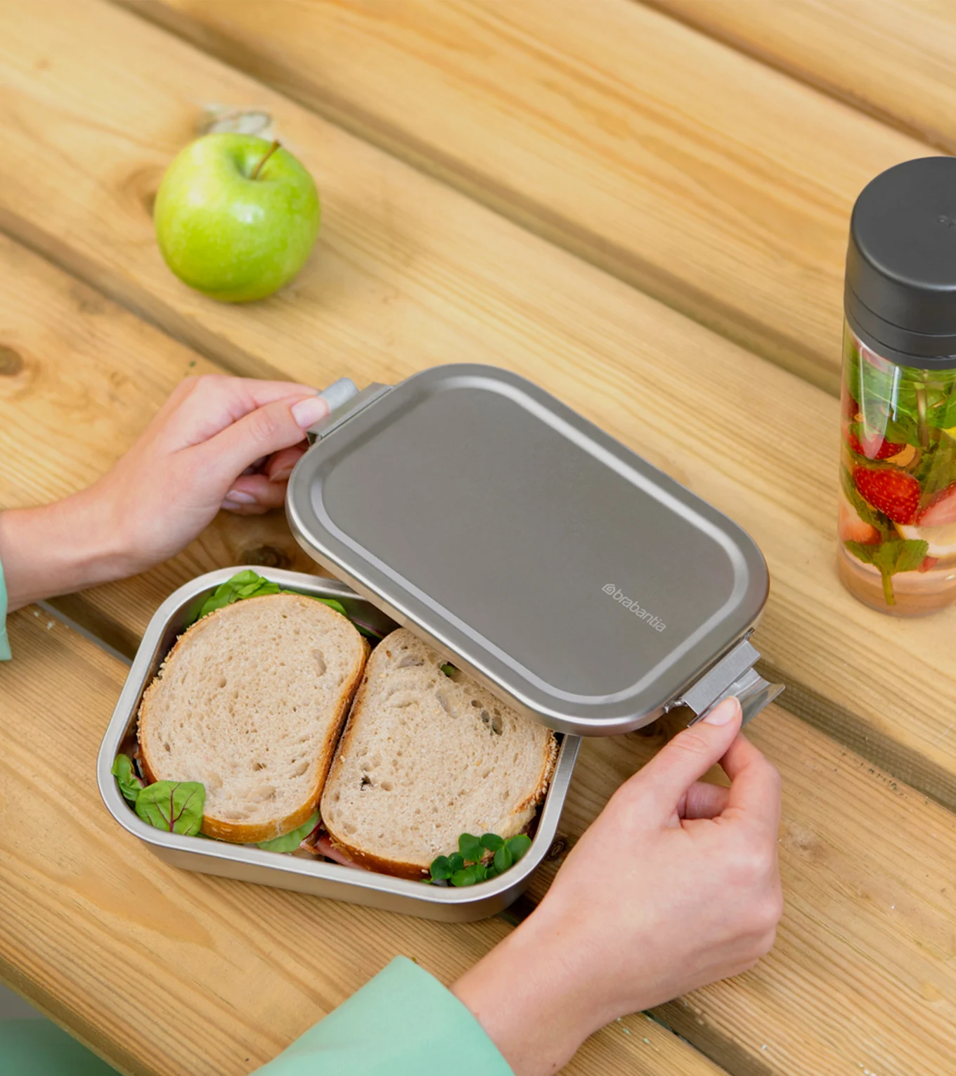 Hände öffnen eine silberne Brotdose mit Sandwiches, einem Apfel und einer Wasserflasche mit Früchten auf einem Holztisch.