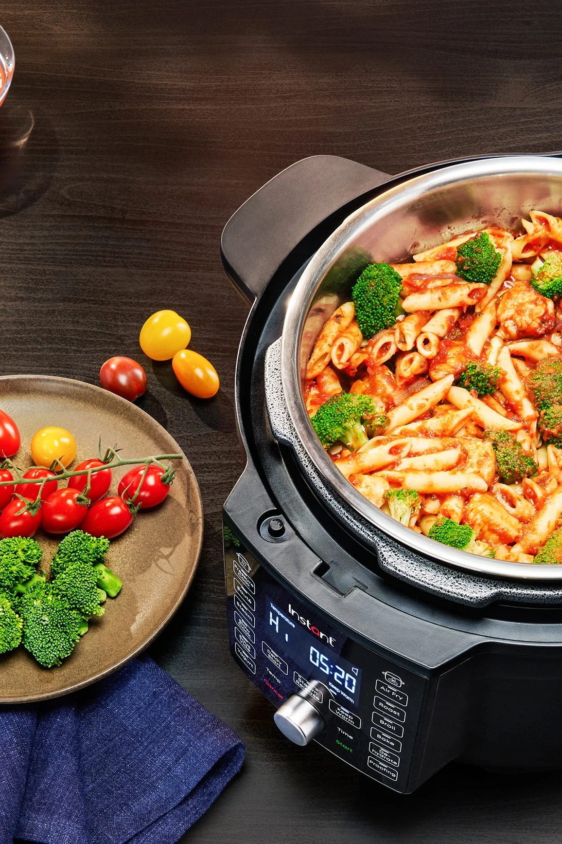 Ein schwarzer Multikocher gefüllt mit Penne-Nudeln und Brokkoli in Tomatensauce, daneben ein Teller mit frischen Kirschtomaten und Brokkoli auf einem dunklen Holztisch.